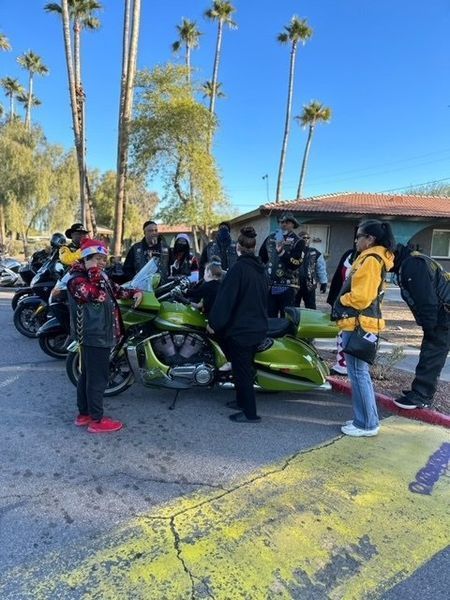 People gather near parked motorcycles; a bright green one stands out. Sunny day, palm trees in background.