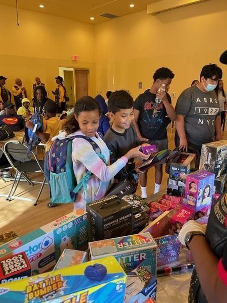 Children selecting toys from a table in a brightly lit room. Other people observe.