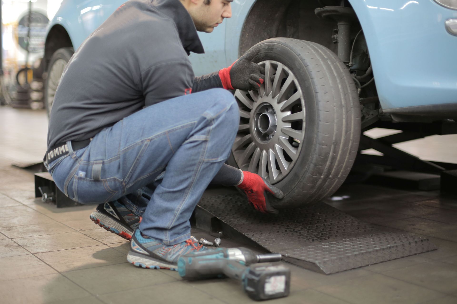 Mechanic kneels beside a car, inspecting a front wheel with tools on the garage floor.