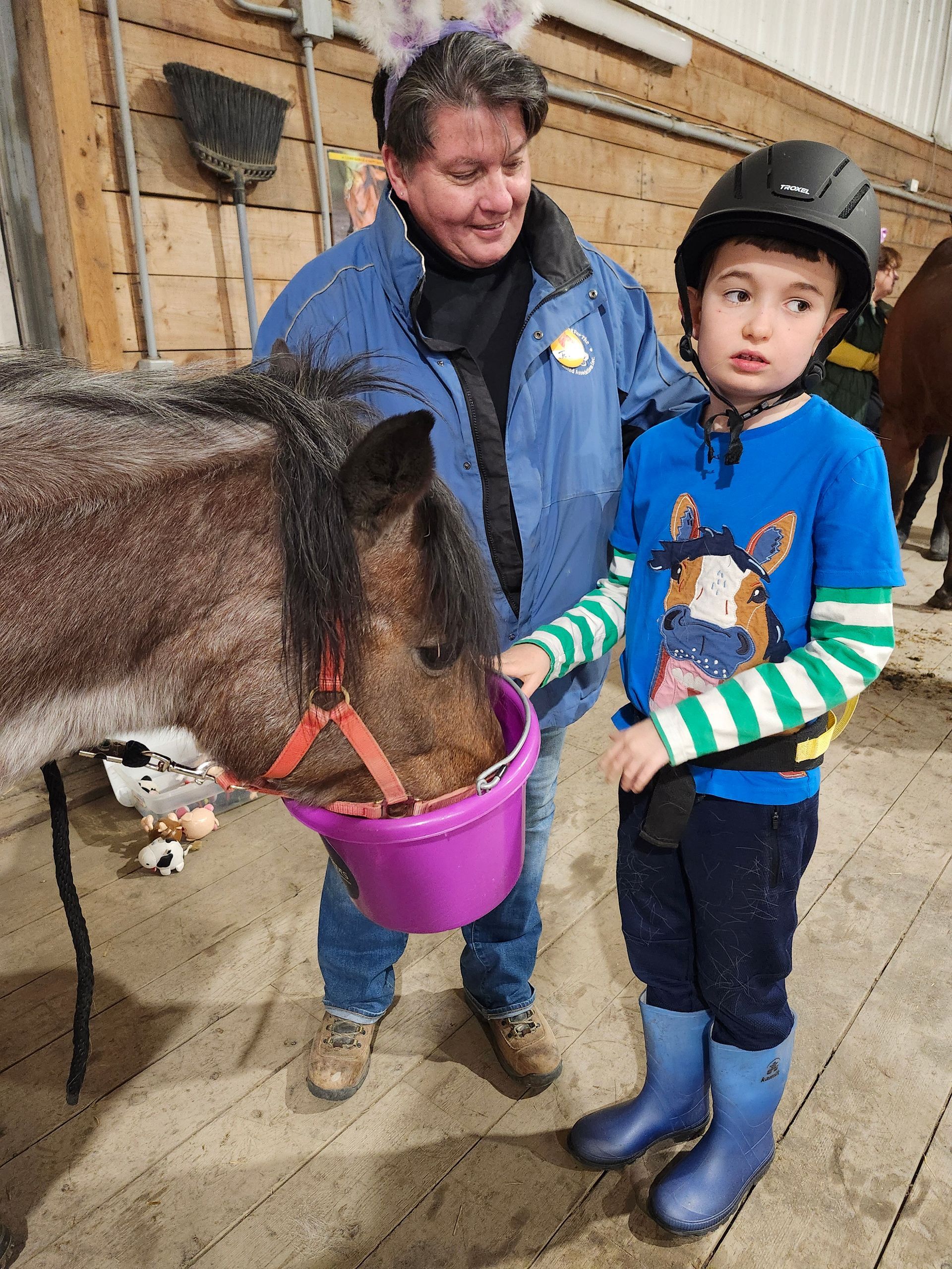 A woman with bunny ears and a young boy feed a horse in a stable. The boy wears a helmet and blue boots.