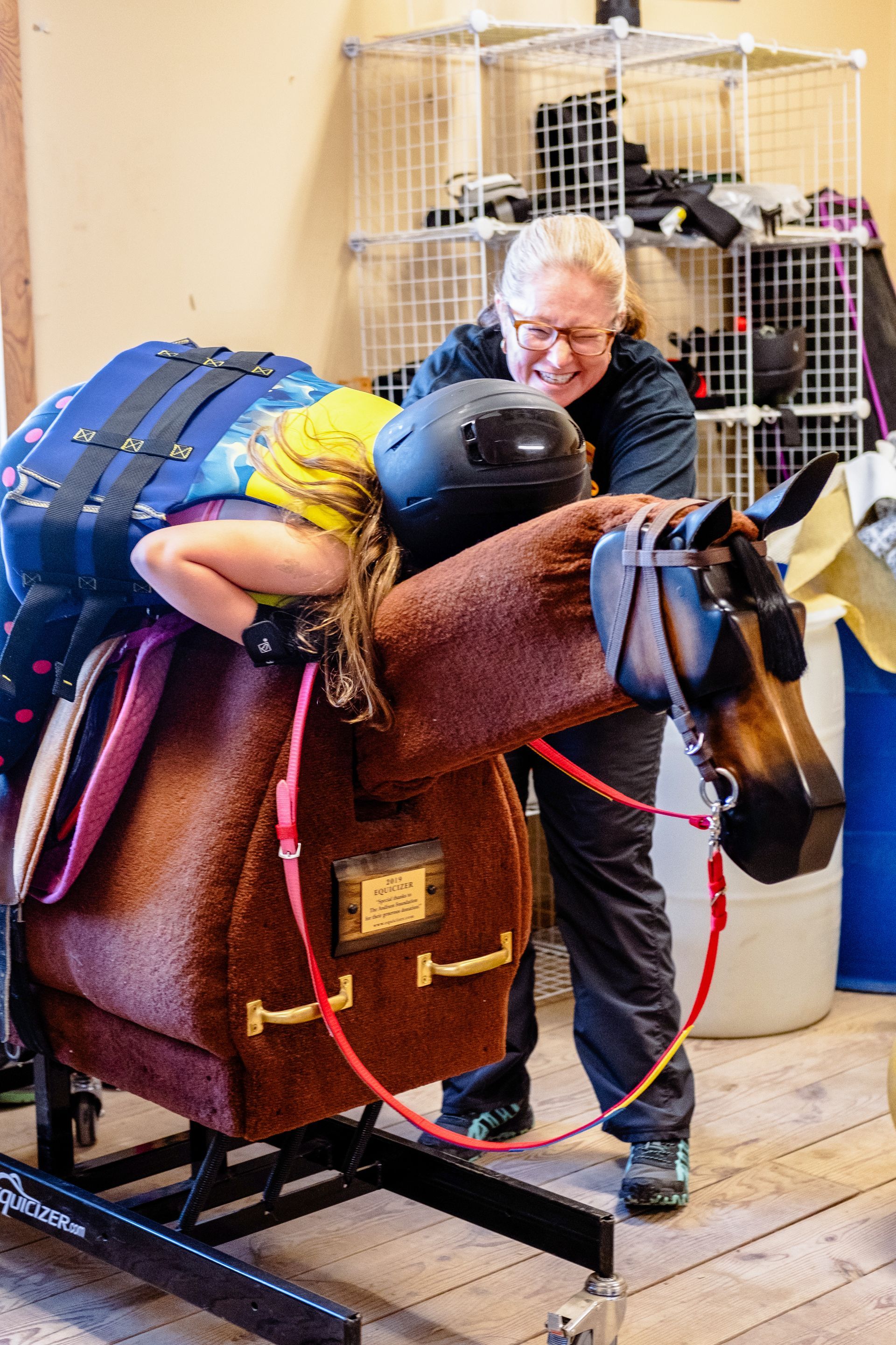 Woman assisting a child with a helmet riding a therapy horse. Both have arms outstretched. Inside setting.