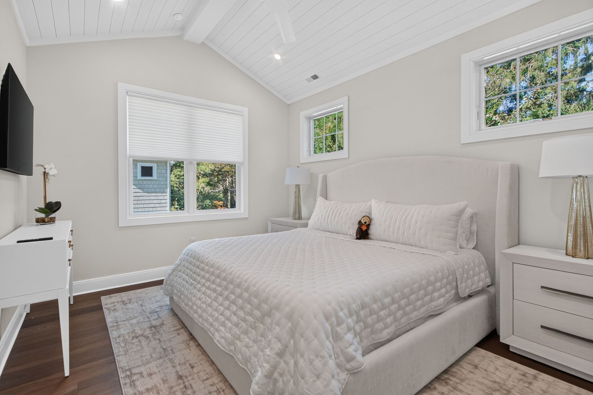 Bright white bedroom with a neatly made bed, windows, dresser, and bedside lamp under vaulted ceiling