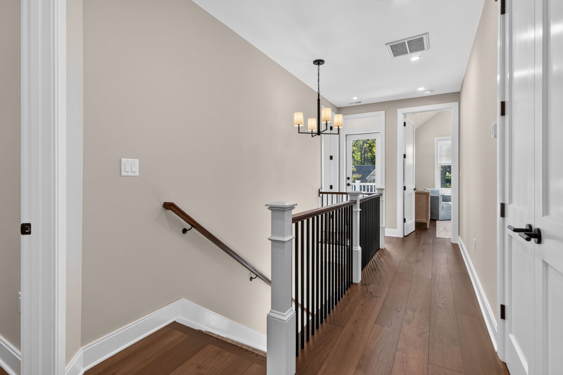 Hallway with wood floors, black stair railing, and pendant light leading to a bright room