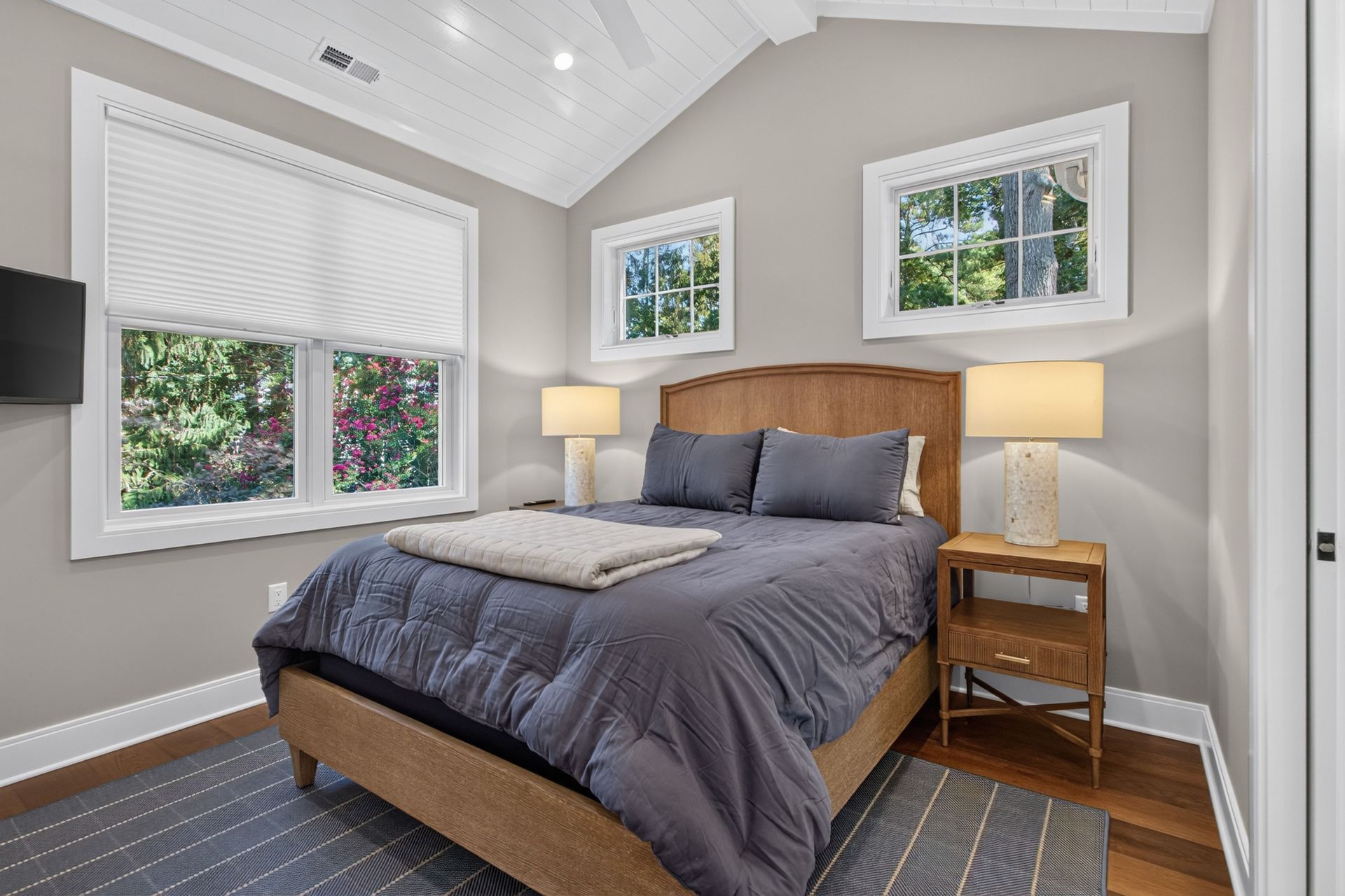 Bright bedroom with a wooden bed, gray bedding, two lamps, and windows on a vaulted ceiling wall