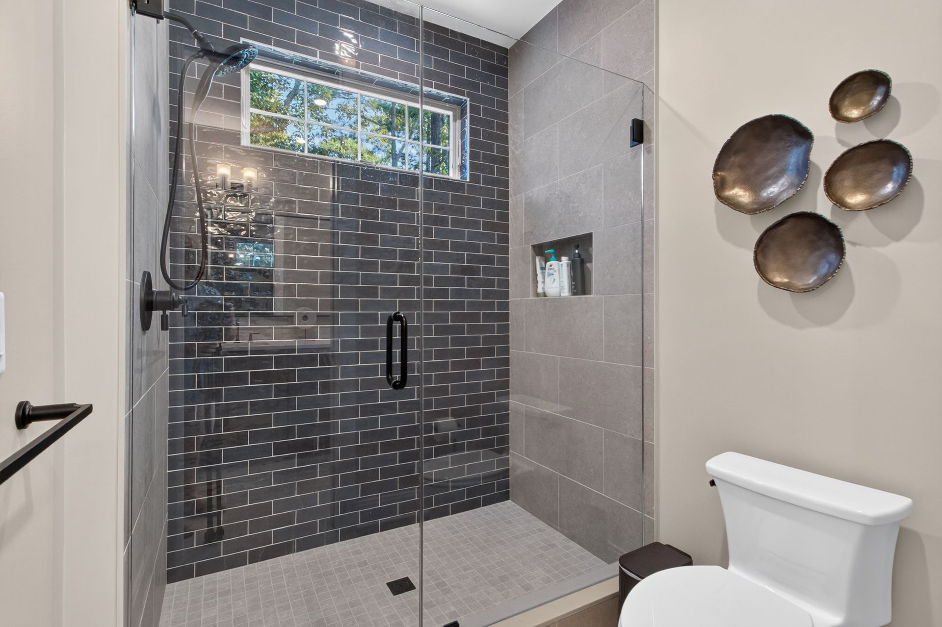 Modern bathroom with glass shower, gray tile, and wall art beside a toilet