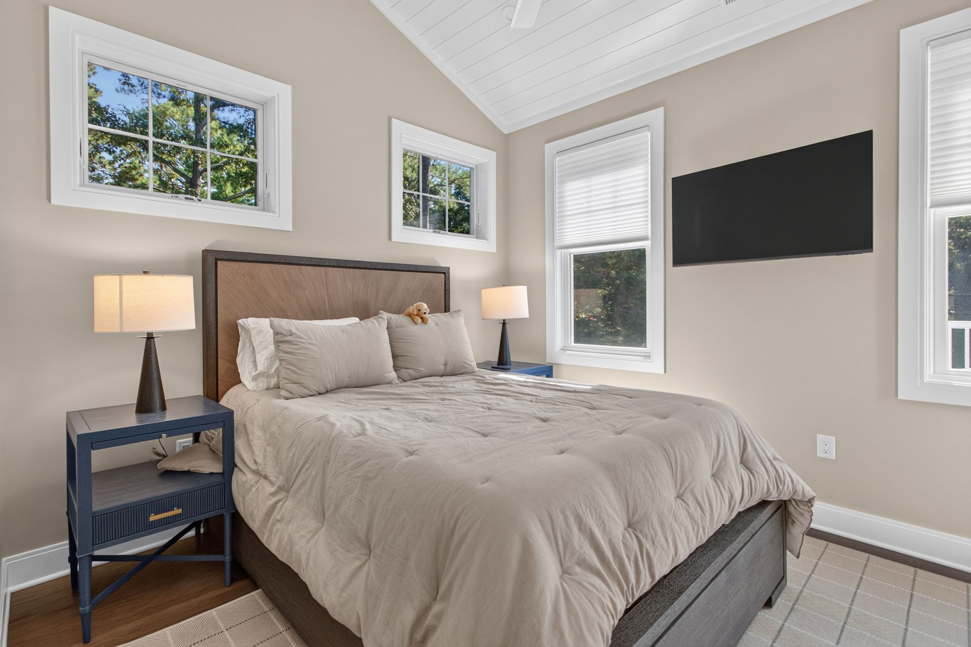 Cozy bedroom with a made bed, bedside lamps, windows, wall art, and a wall-mounted TV