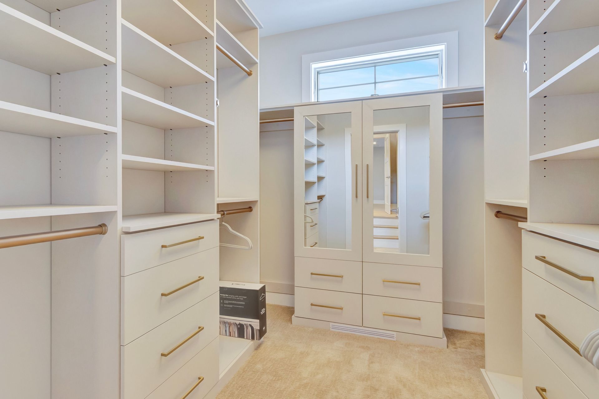 Walk-in closet with white shelving, drawers, and gold hardware. Light-colored carpet and mirrored doors are in the center.