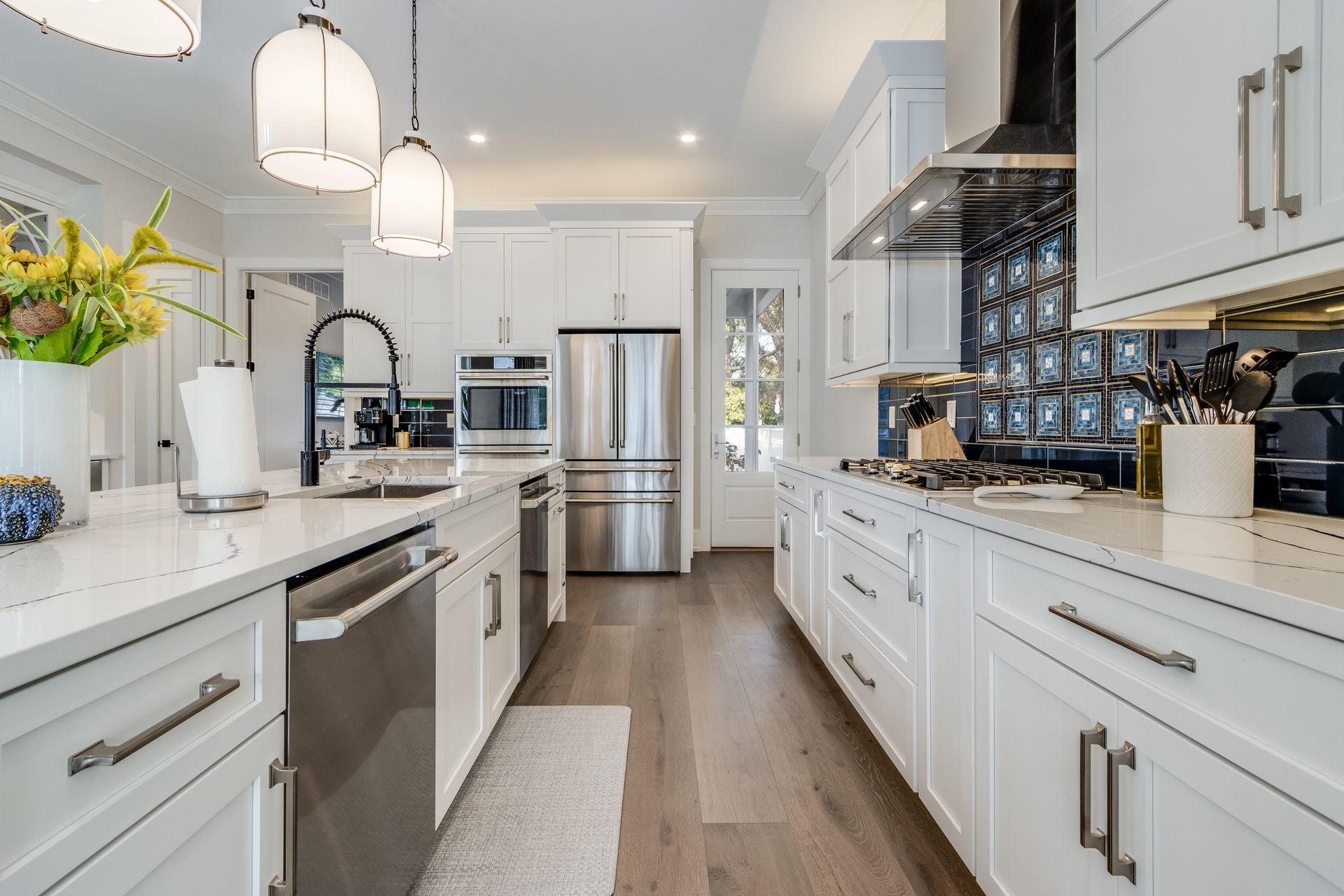 A bright, modern kitchen featuring white shaker cabinets, stainless steel appliances, marble countertops, and wood floors.