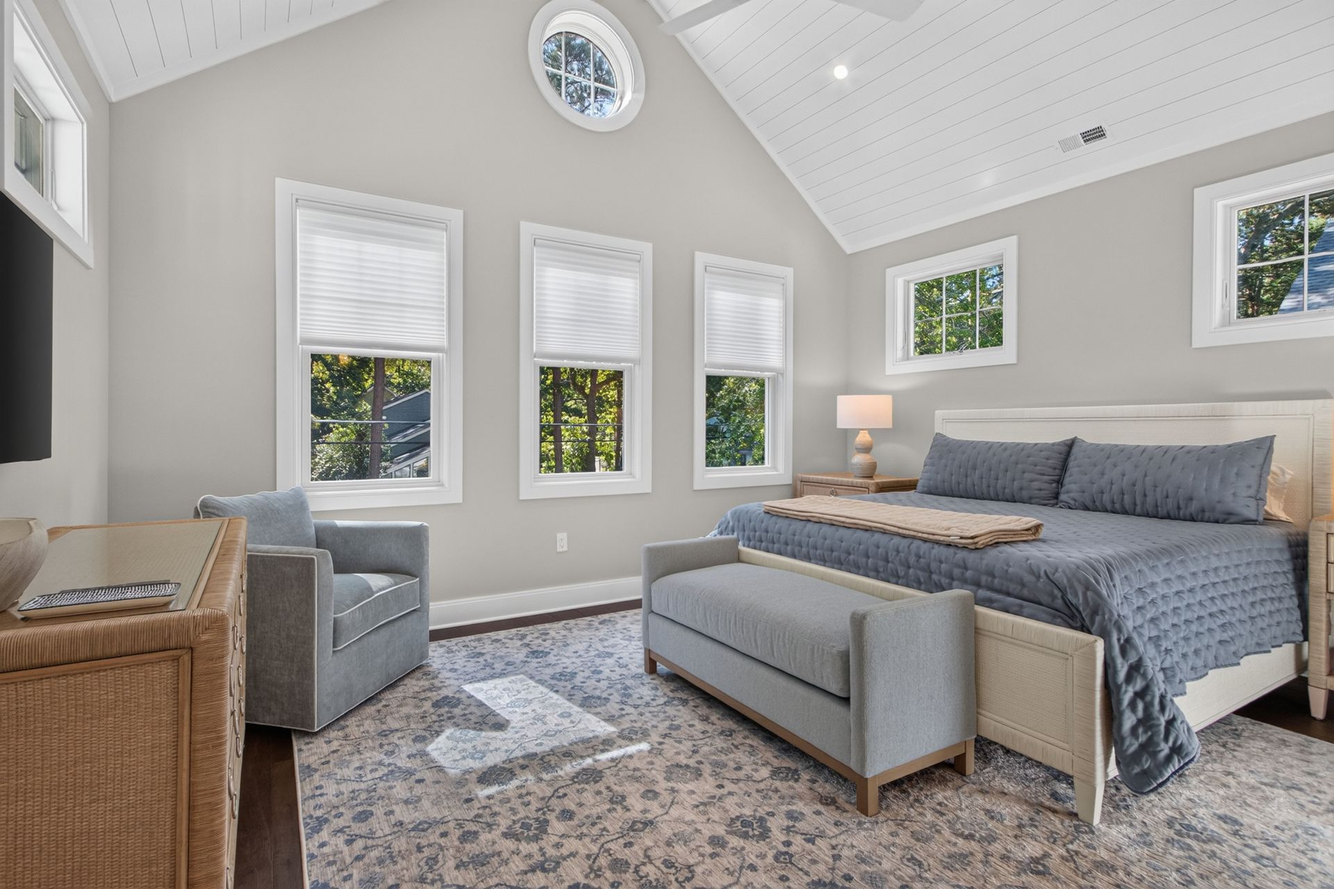 Bright bedroom with gray bed, wicker chair, high vaulted ceiling, and multiple windows under a round wall clock
