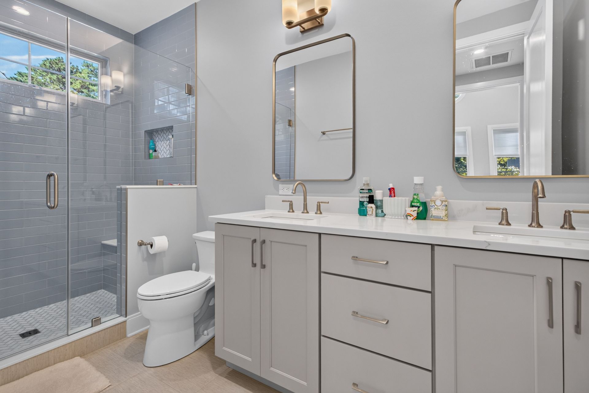 Bright modern bathroom with glass shower, toilet, and double vanity with mirrors and gray cabinetry.