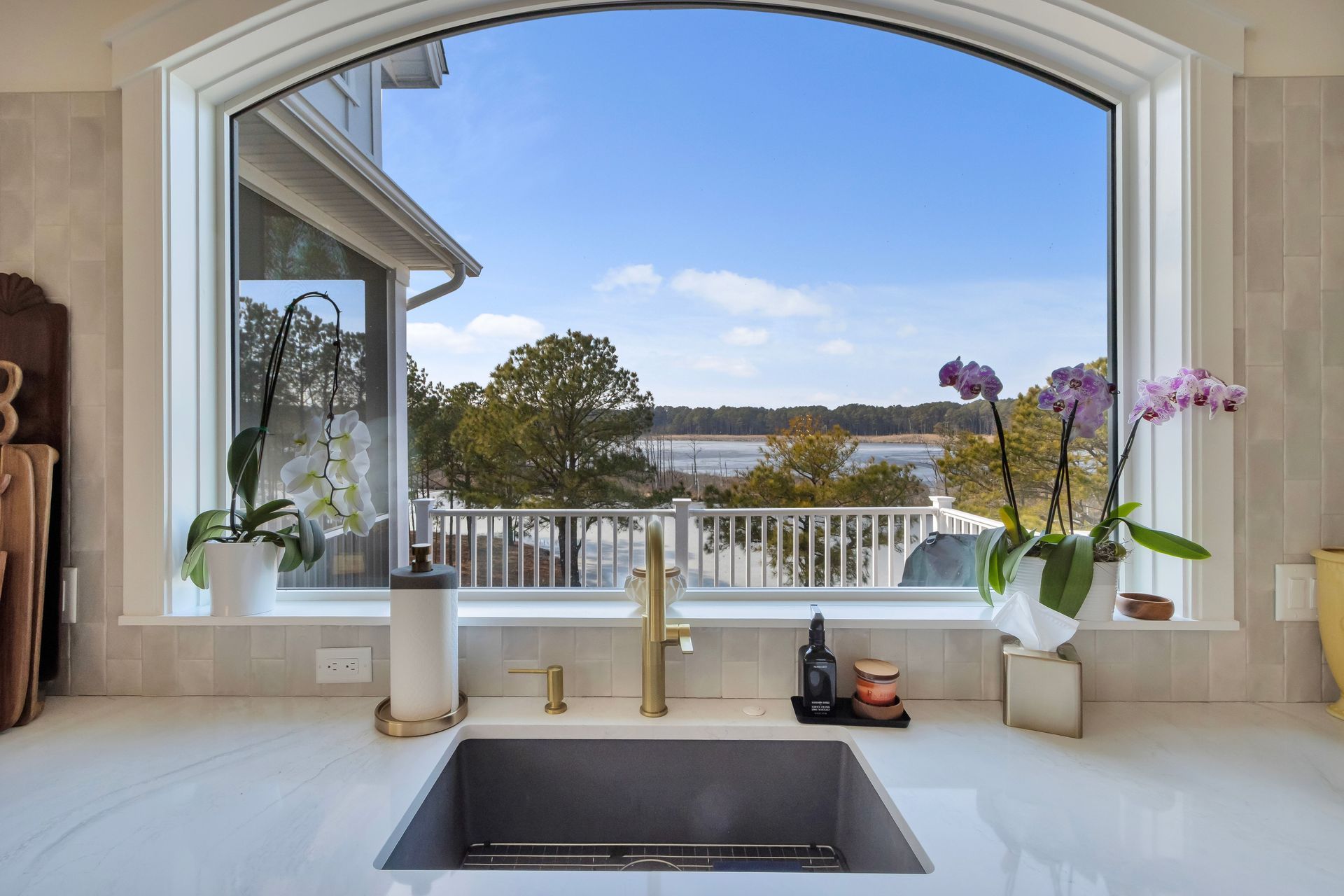 Kitchen sink with arched window overlooking water and trees, gold faucet and orchids on countertop.