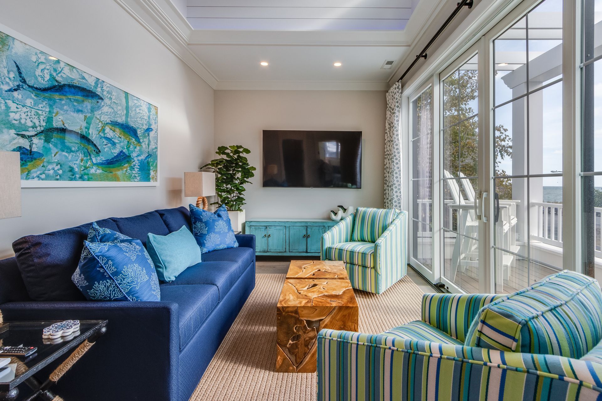 Coastal-themed living room with blue sofa, striped chairs, TV, artwork of fish, and balcony access.