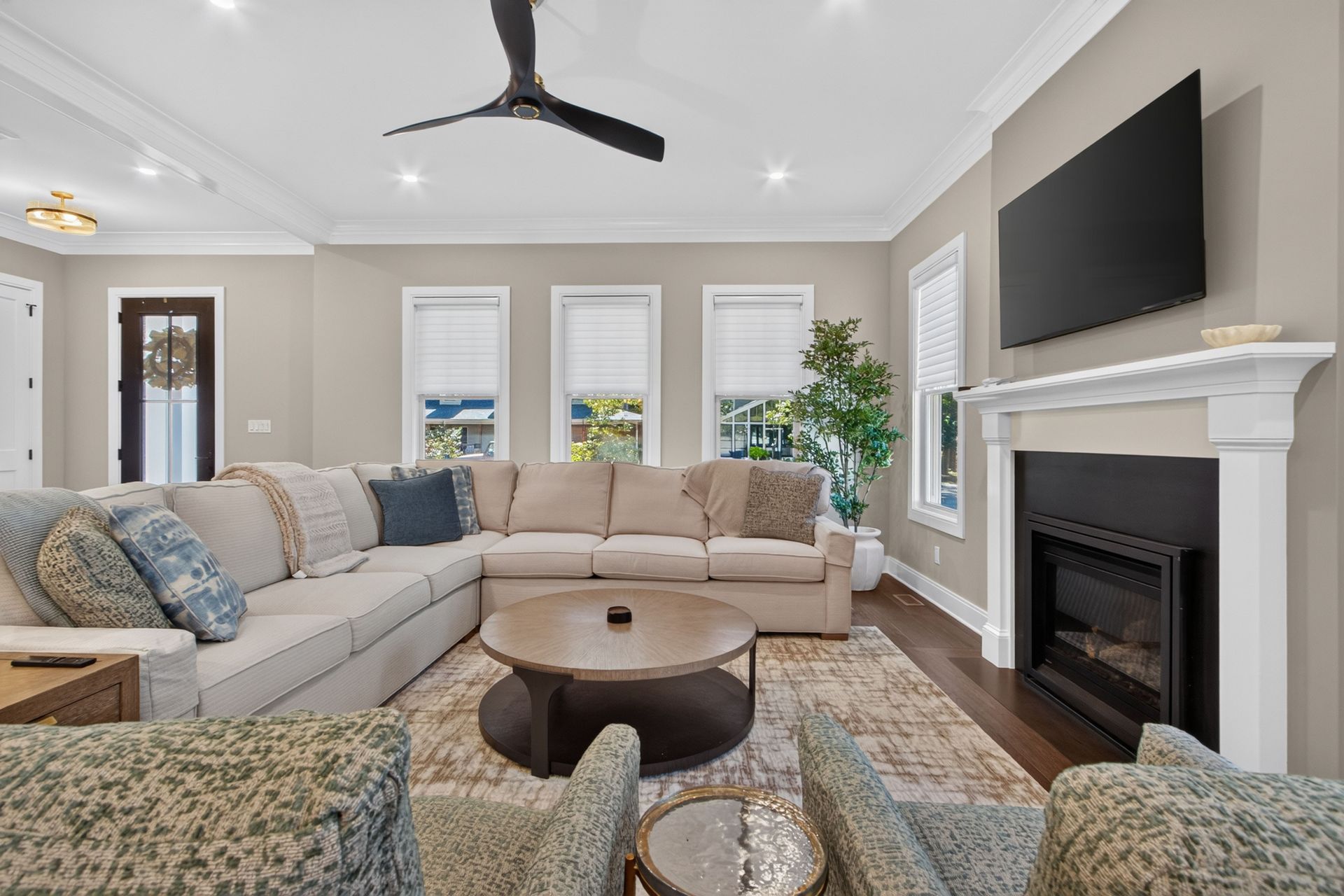 Bright living room with beige sectional, patterned chairs, round coffee table, fireplace, and wall-mounted TV.