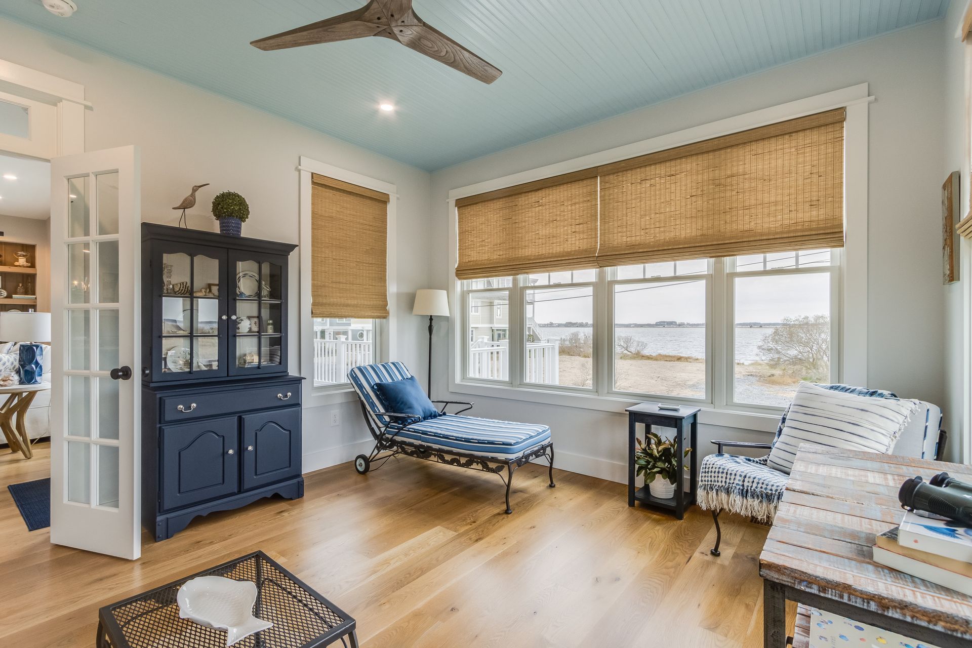 Sunroom with blue ceiling, wood floors, and water view. Furnished with a chaise lounge, armchair, and cabinet.