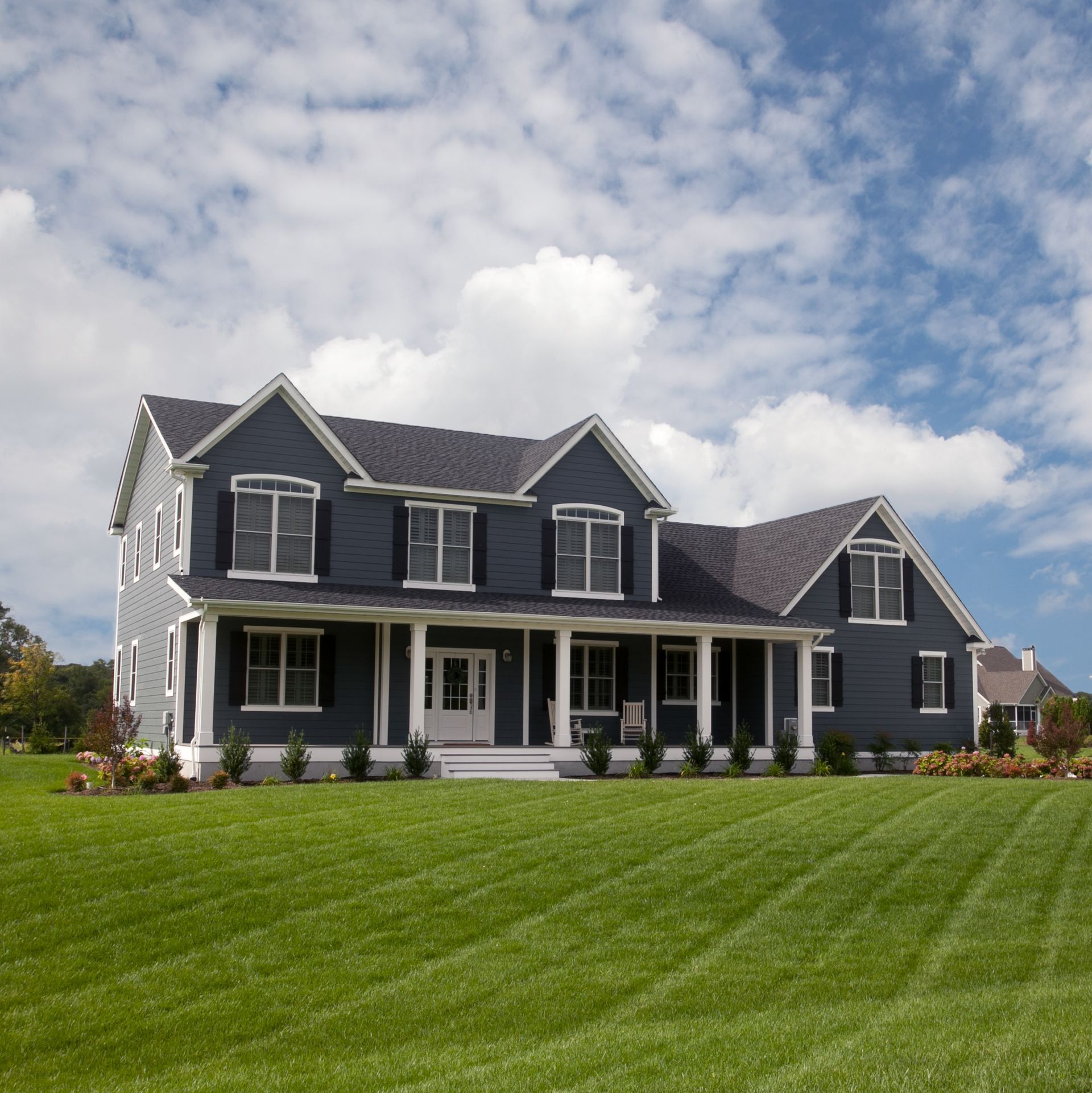 Large dark-gray suburban house with a white porch on a wide green lawn under a partly cloudy sky