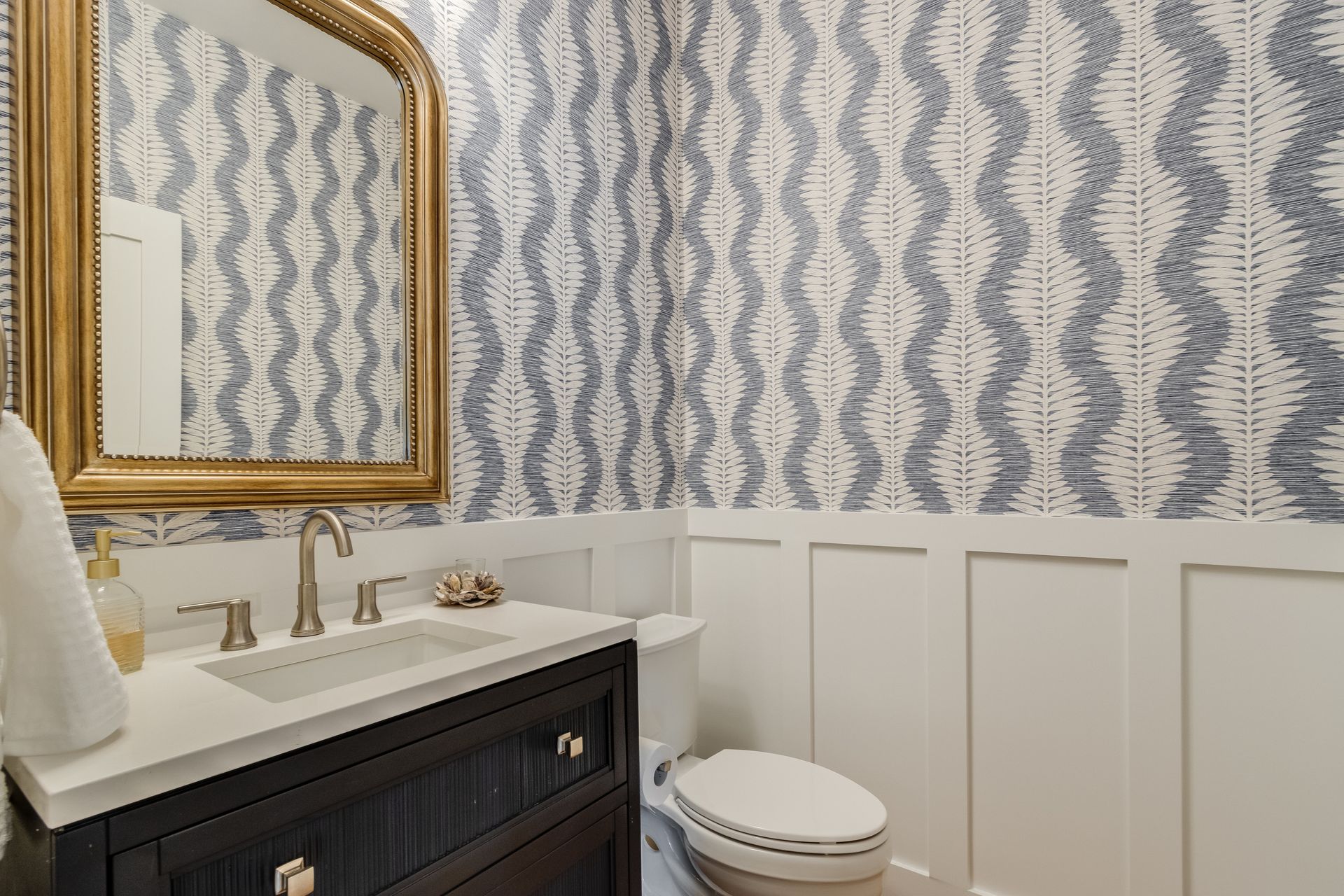 Bathroom with gold mirror, blue and white patterned wallpaper, white wainscoting, and dark vanity.