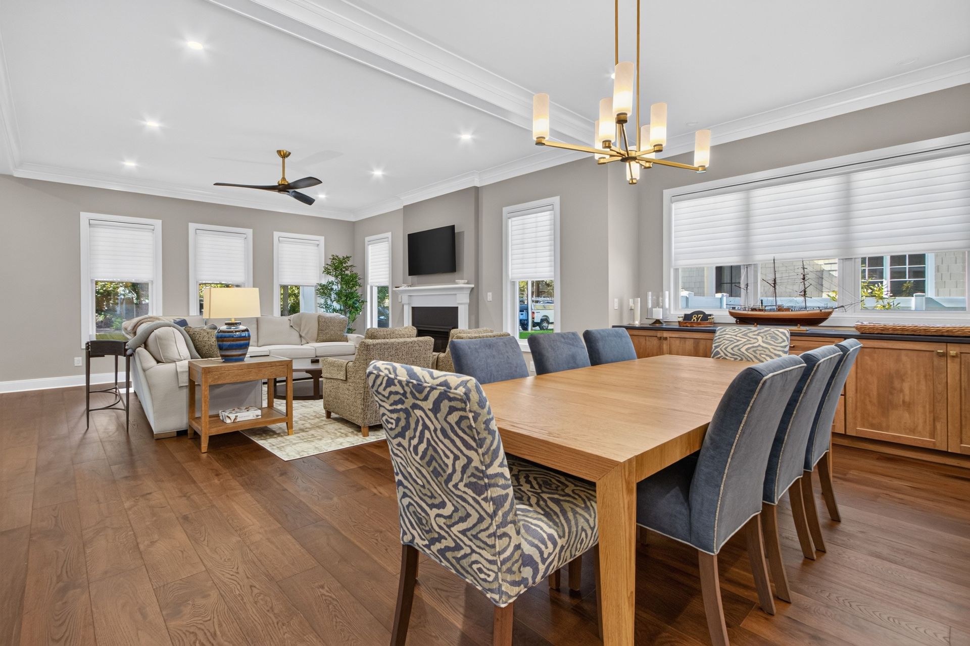 Bright open-concept living and dining room with wood floors, gray sofas, and a kitchen in the background