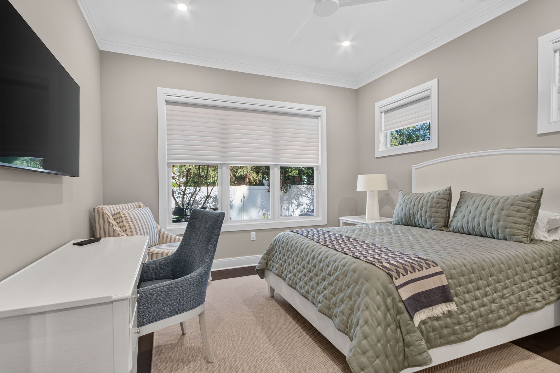 Bright bedroom with bed, desk, chair, wall-mounted TV, and window with blinds