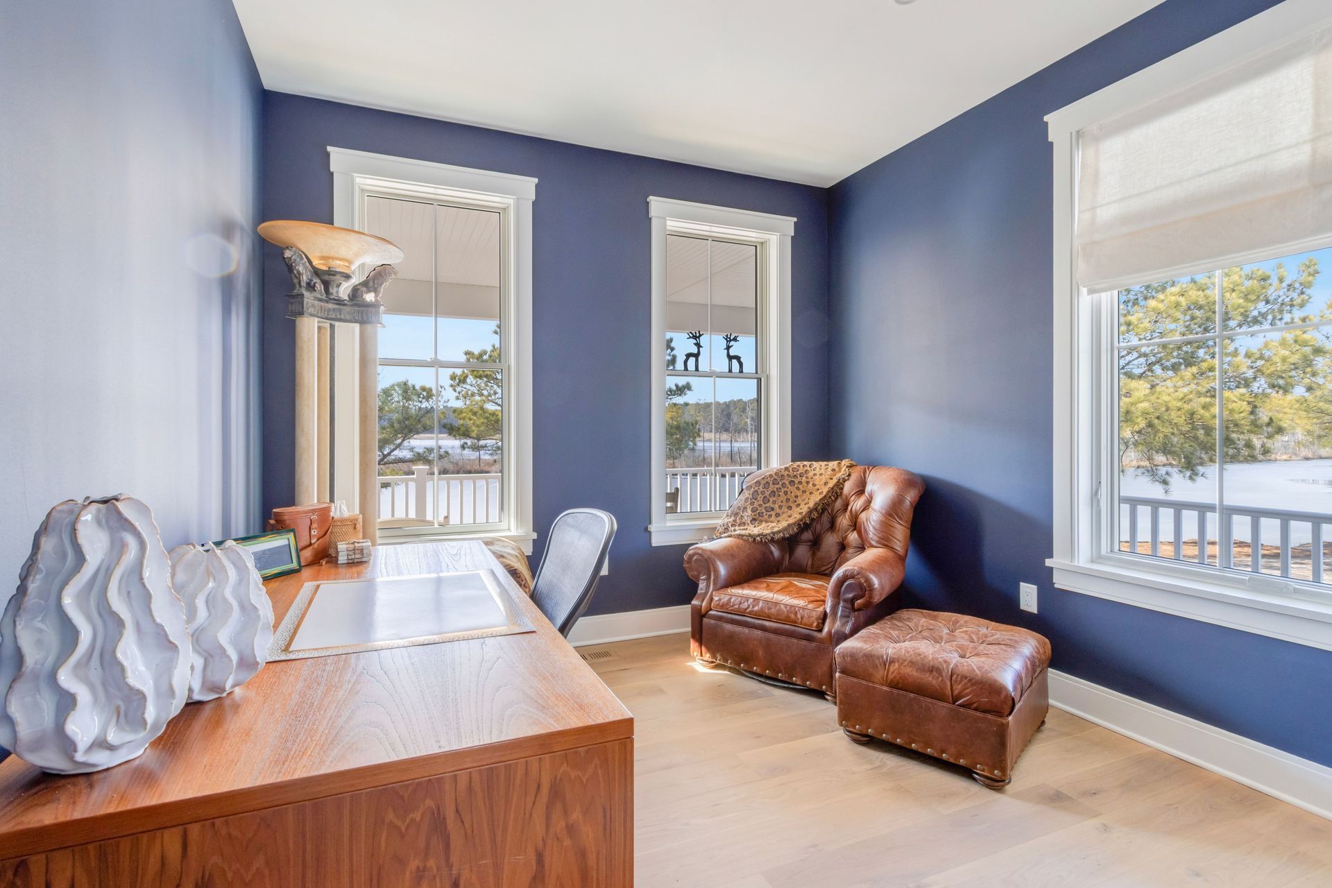 Blue-walled home office with wood desk, leather chair, and windows overlooking a porch and water.
