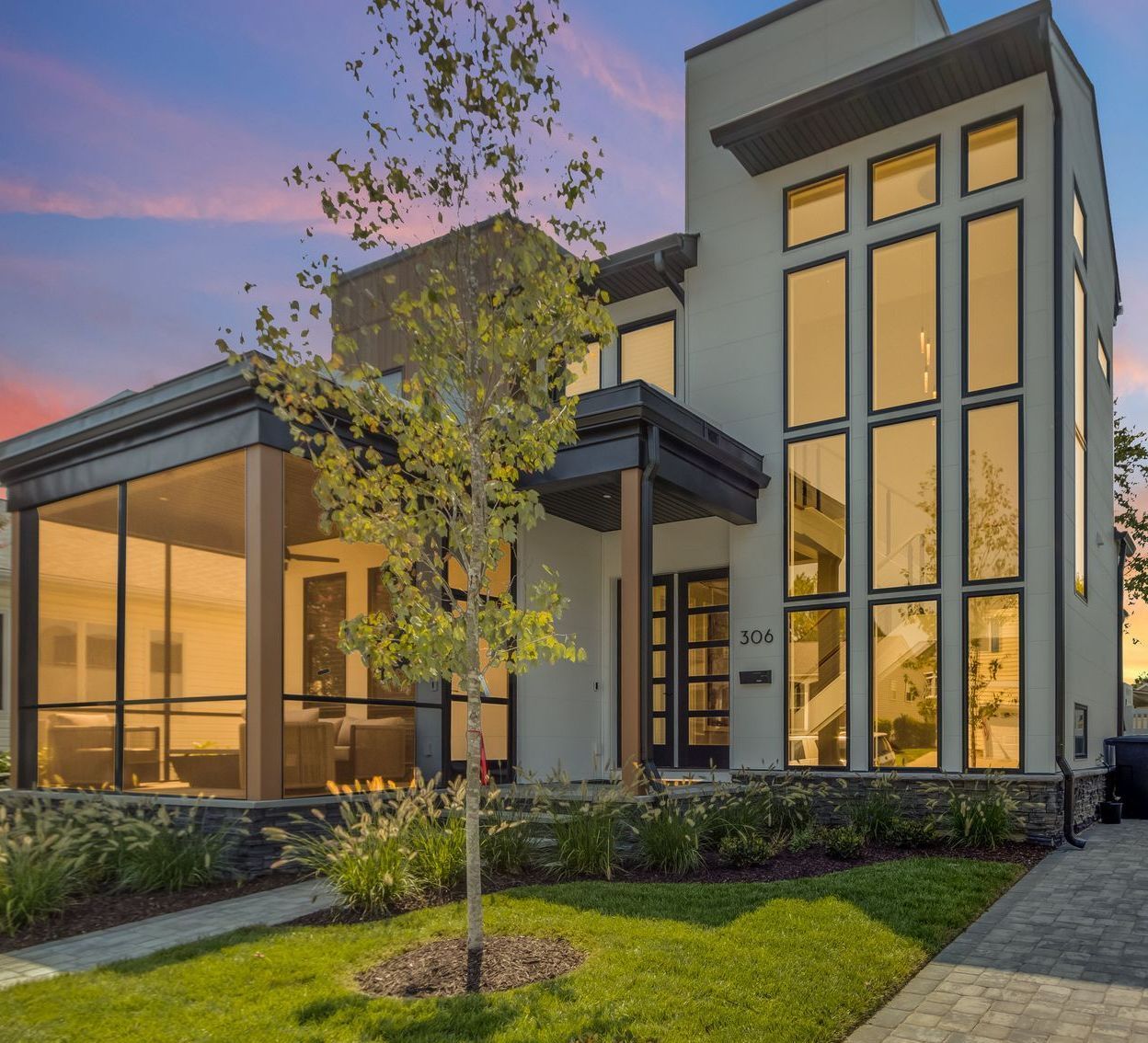 Modern house with large windows, a screened porch, and a tree in front at dusk.