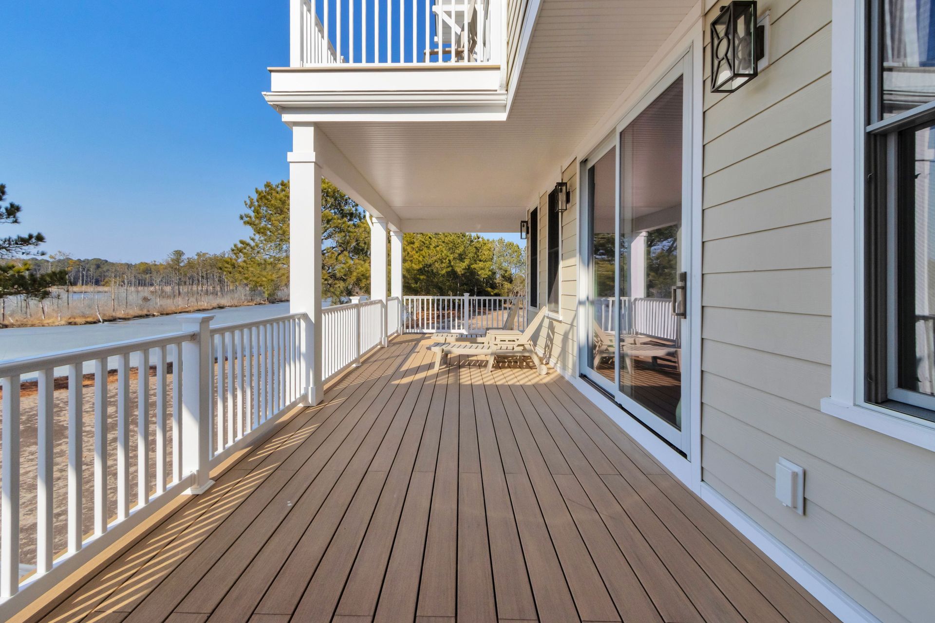 A white porch with light brown wood decking overlooks a waterway. Glass sliding doors and white railings are visible.