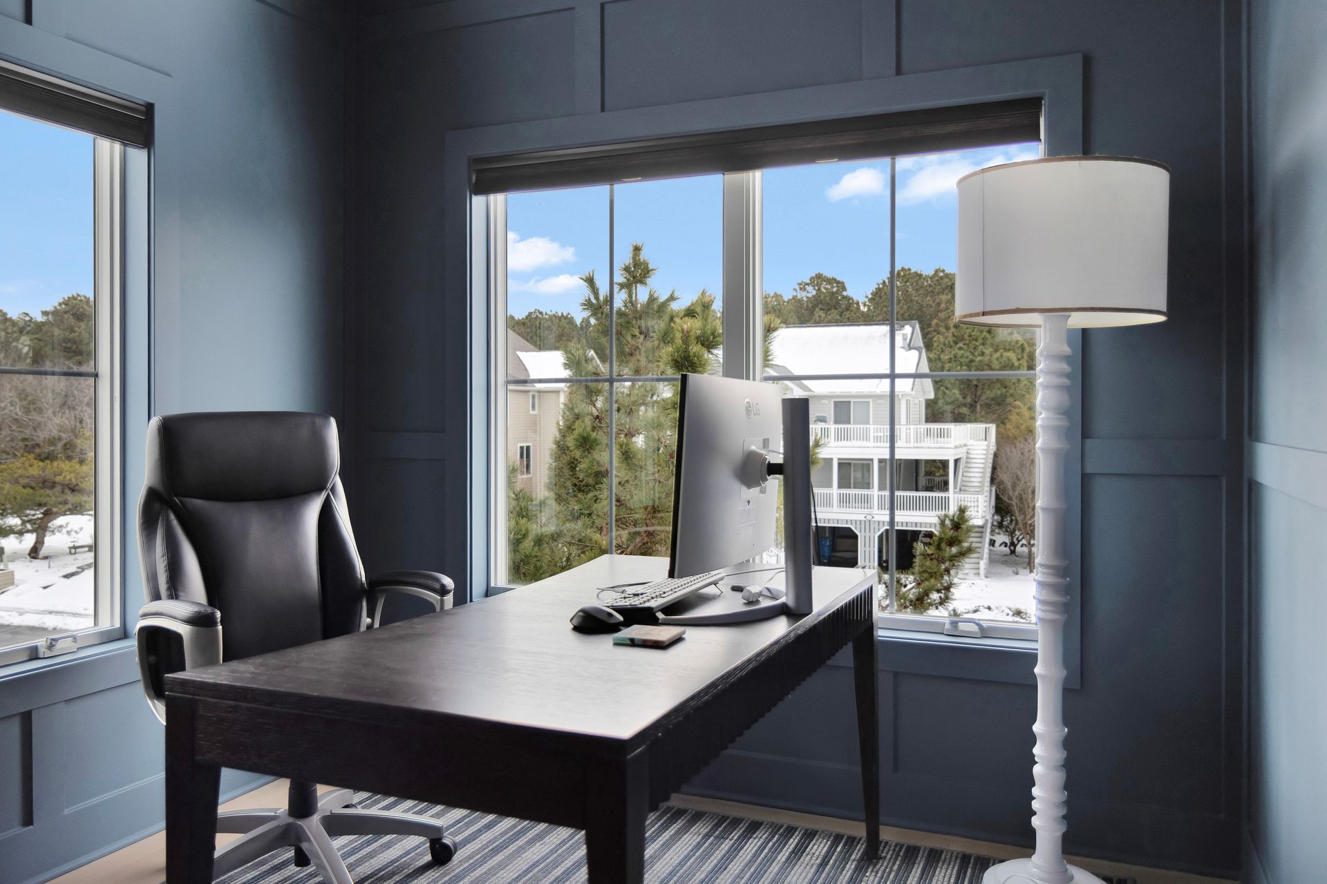 Home office with black desk, chair, and computer, looking out at snowy rooftops through a large window. Blue walls.