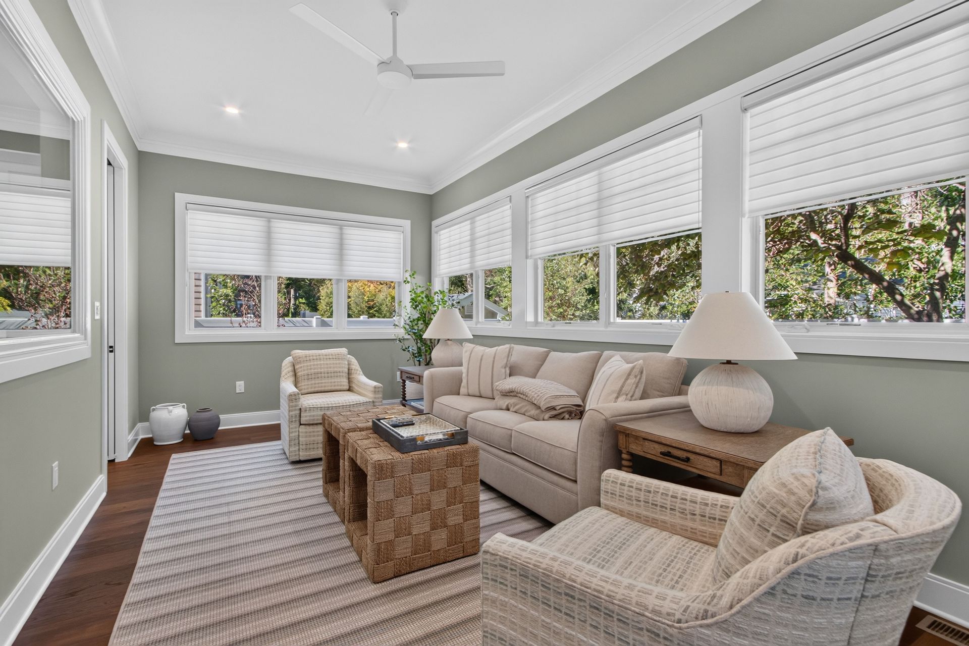Bright sunroom with beige sofas, woven chairs, striped rug, and large windows with white blinds.