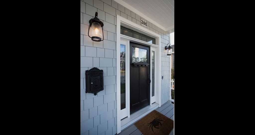 Black front door with sidelights and mailbox on light blue shingle siding; outdoor lamp.