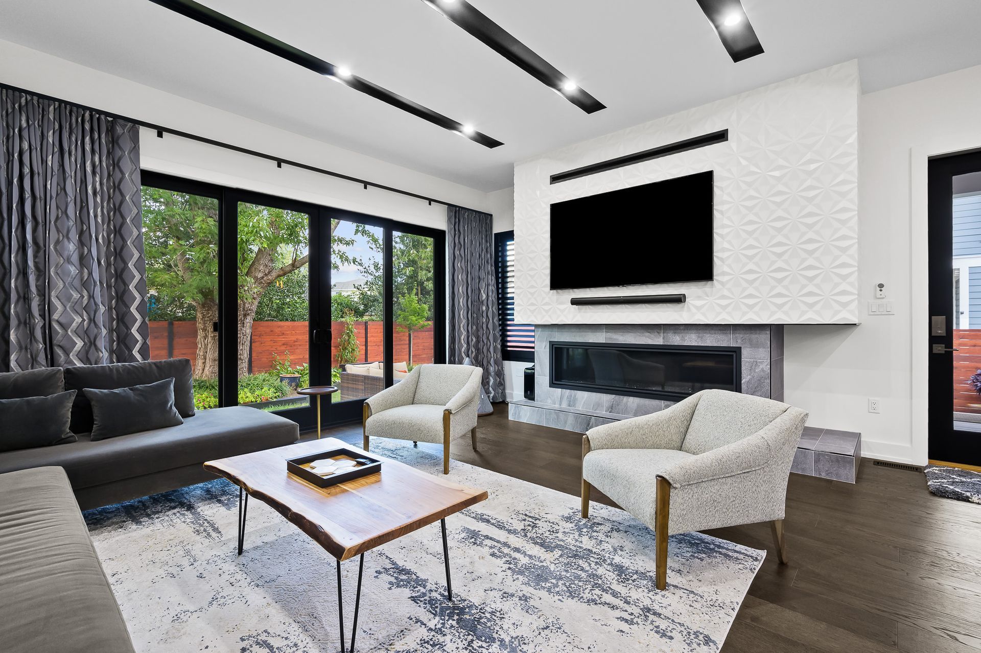 Living room with gray sofa, patterned rug, white chairs, and fireplace beneath a large TV.