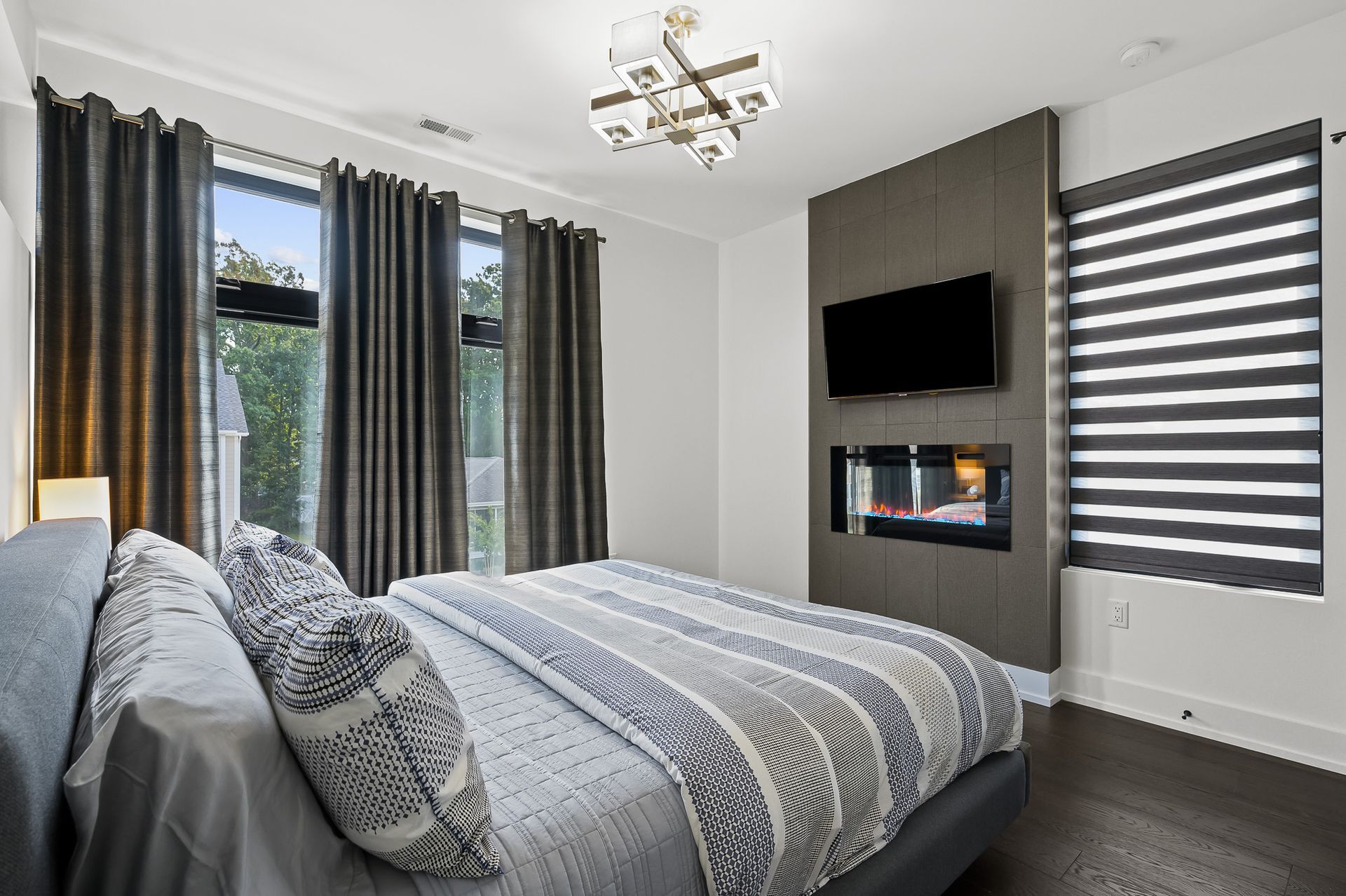 Bedroom with a bed, fireplace, TV, and large windows with gray curtains and blinds.