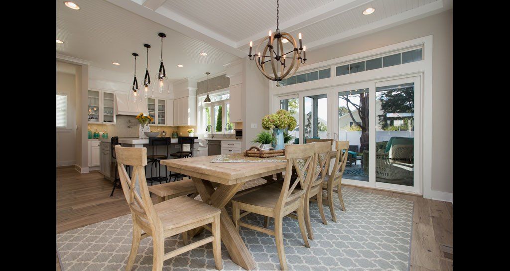 Dining room with a wooden table, chairs, and chandelier. Kitchen in the background. Glass doors to the outside.