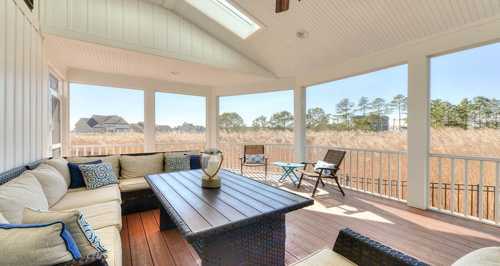 Covered porch with sectional sofa, table, and chairs overlooking marsh and trees.