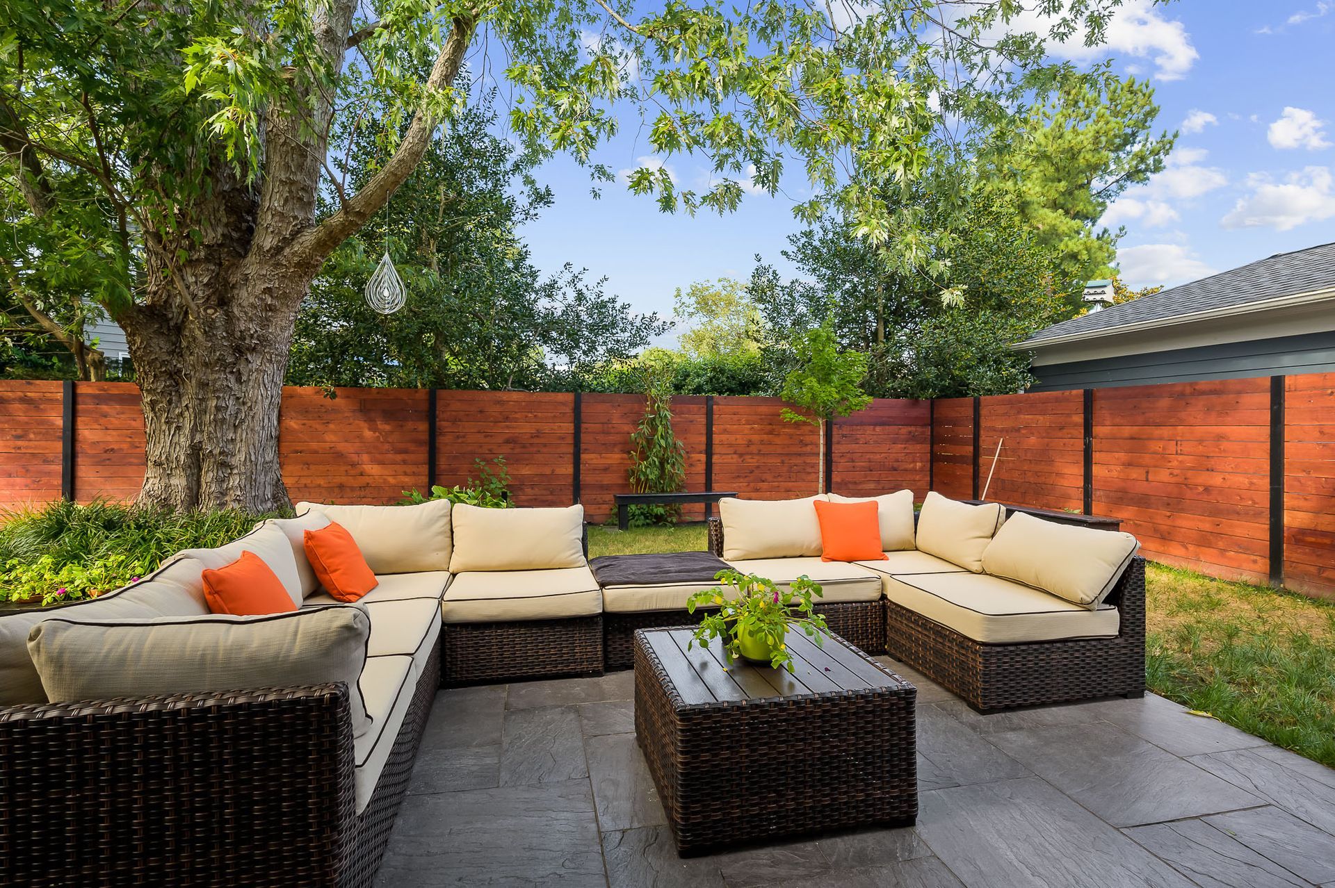 Patio with brown wicker furniture, orange pillows, and a wooden fence under a blue sky.