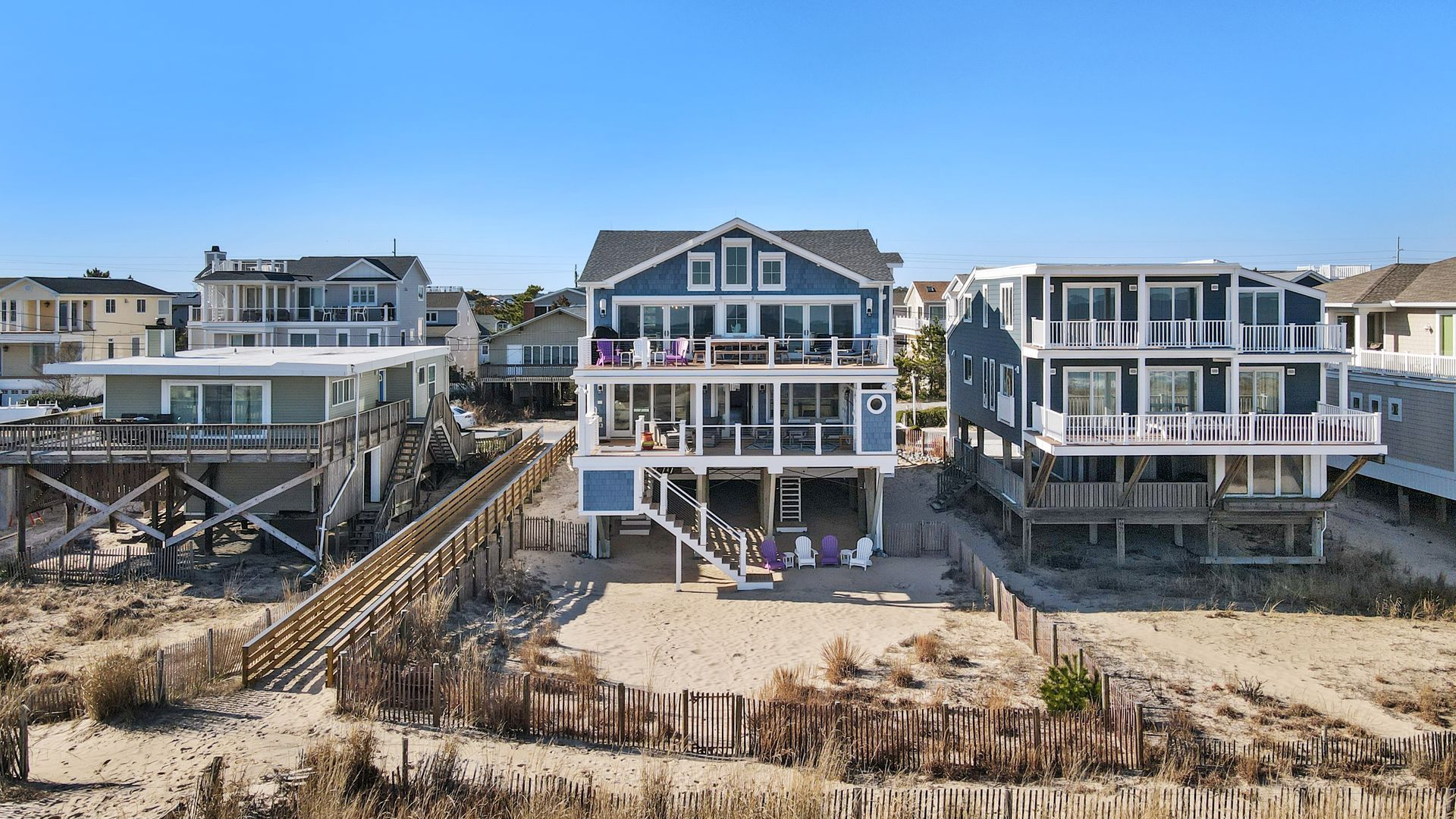Beachfront homes with decks and stairs, blue and grey exteriors, sand dunes in foreground.