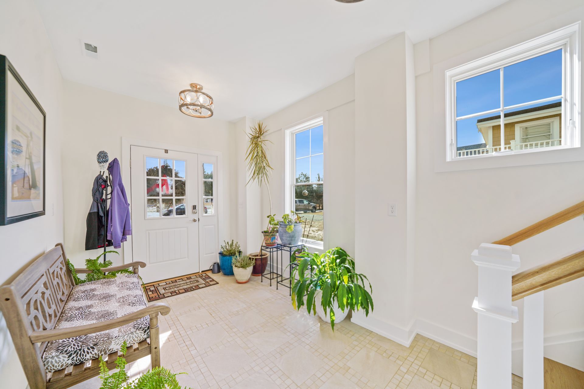 Bright entryway with a bench, potted plants, white walls, and a staircase by a sunlit window.