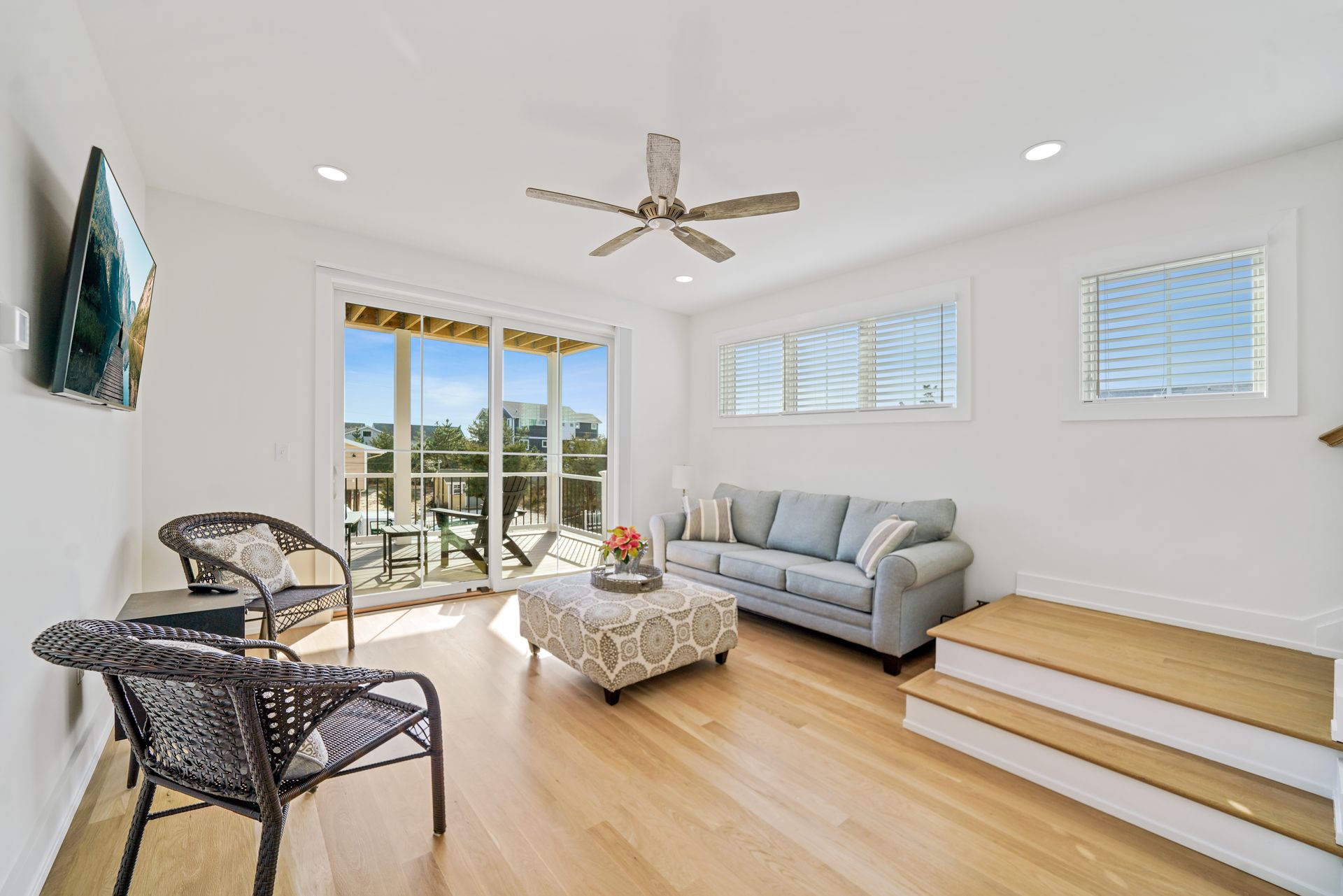 Bright living room with wicker chairs, gray sofa, white steps, ceiling fan, and open glass doors to a balcony.