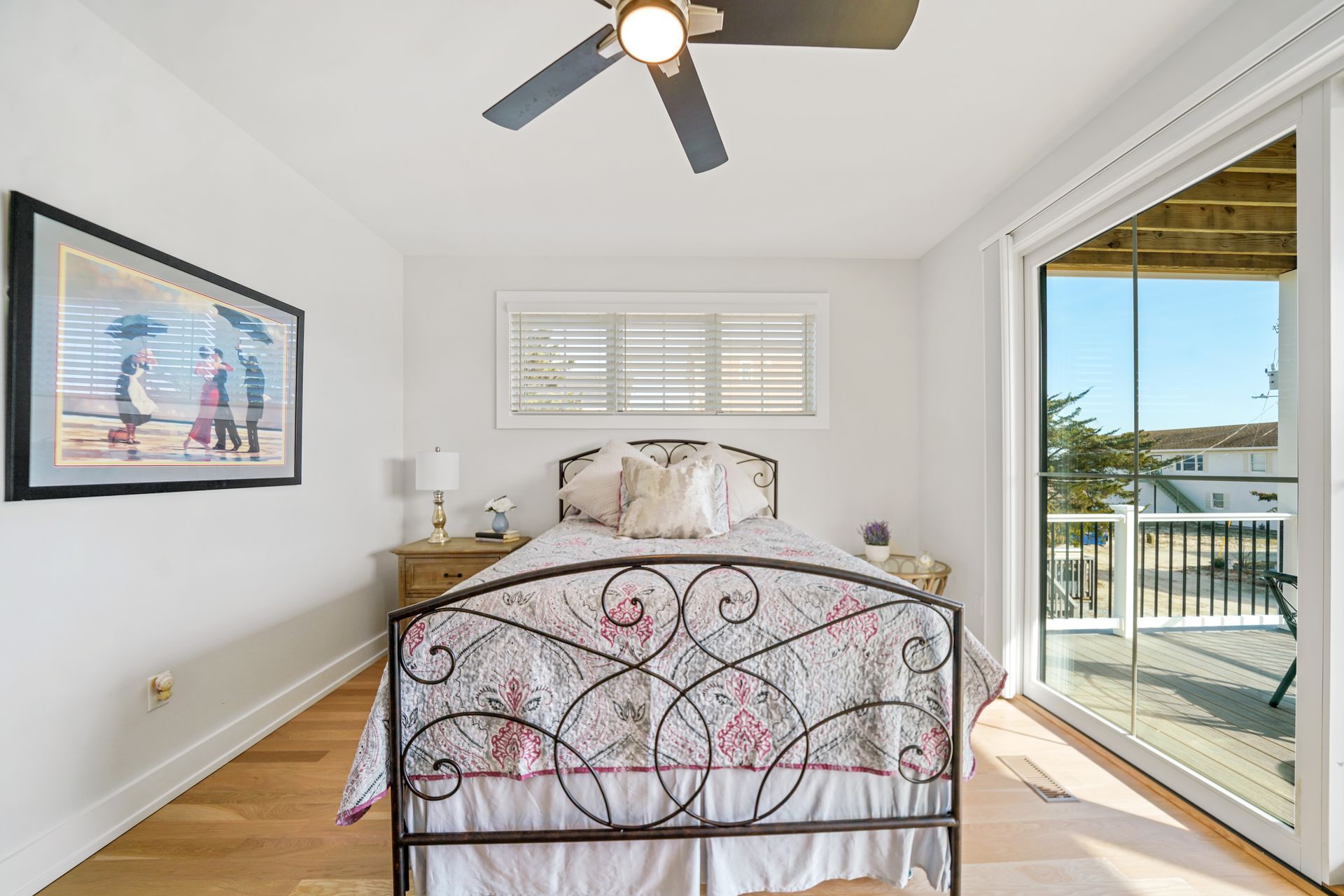 Bright bedroom with metal bed, beach artwork, ceiling fan, and sliding glass door to a balcony