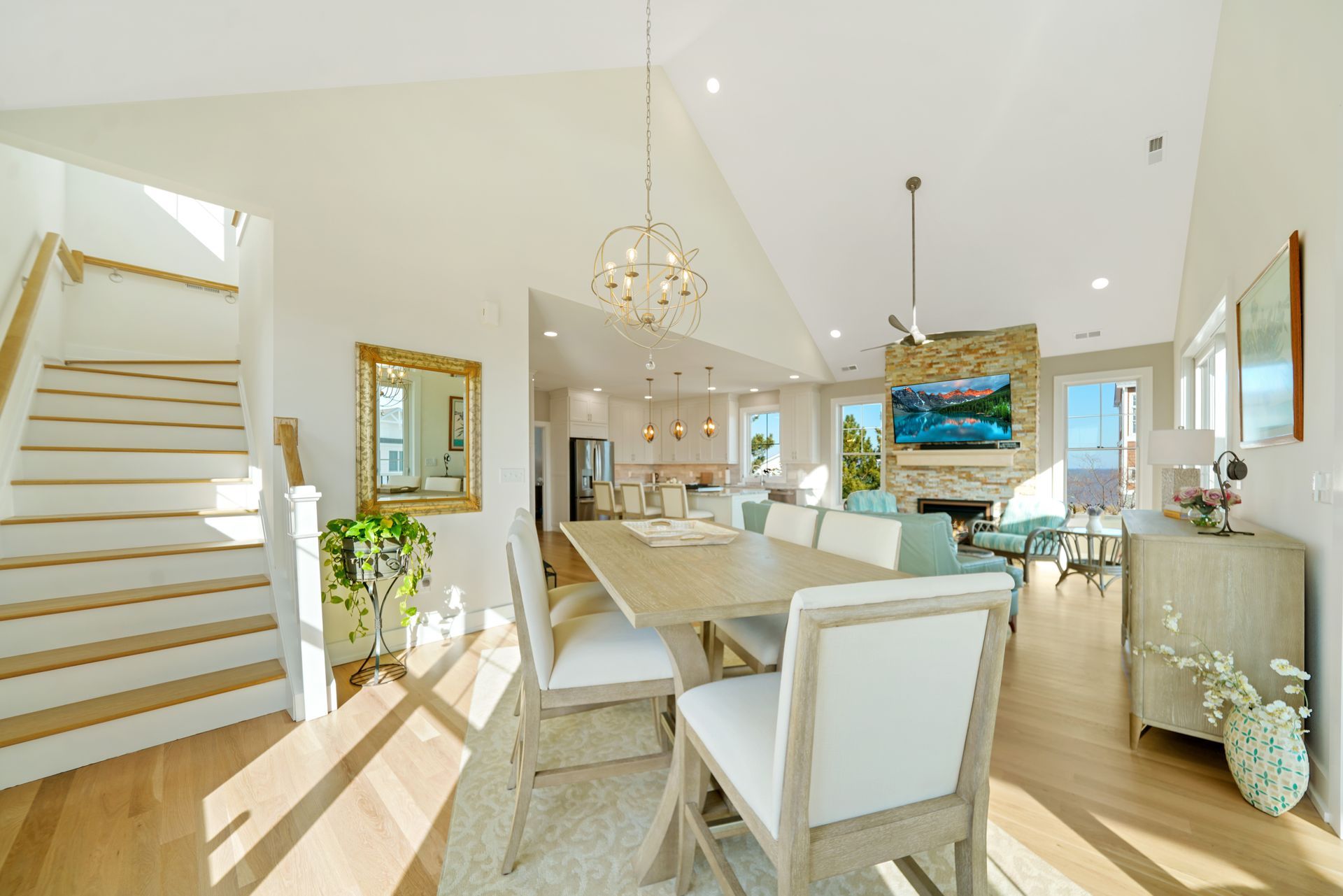 Bright open dining room with white chairs, wooden table, staircase, and large windows overlooking a scenic view