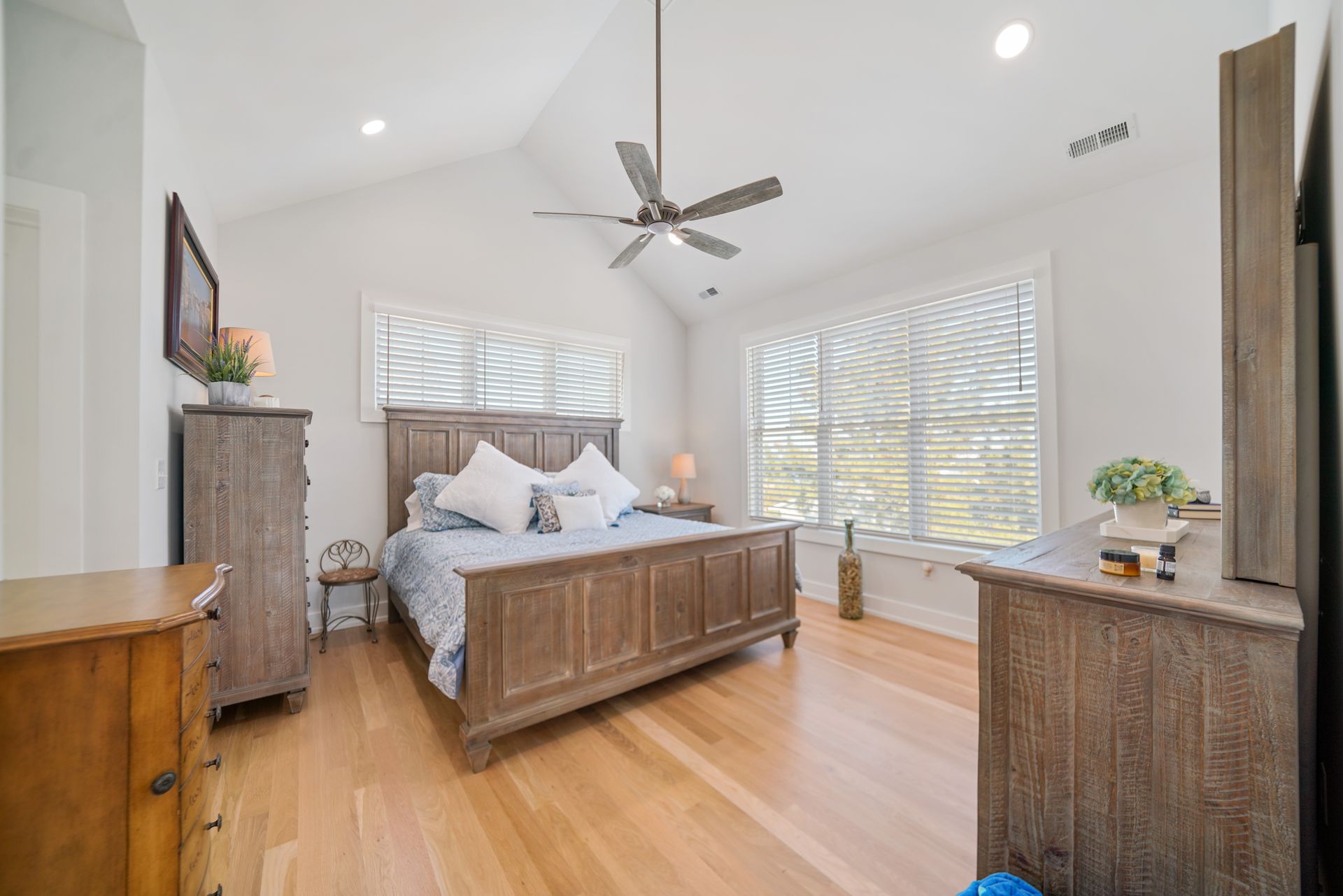 Bright bedroom with a wooden bed, dresser, ceiling fan, and large windows letting in daylight.