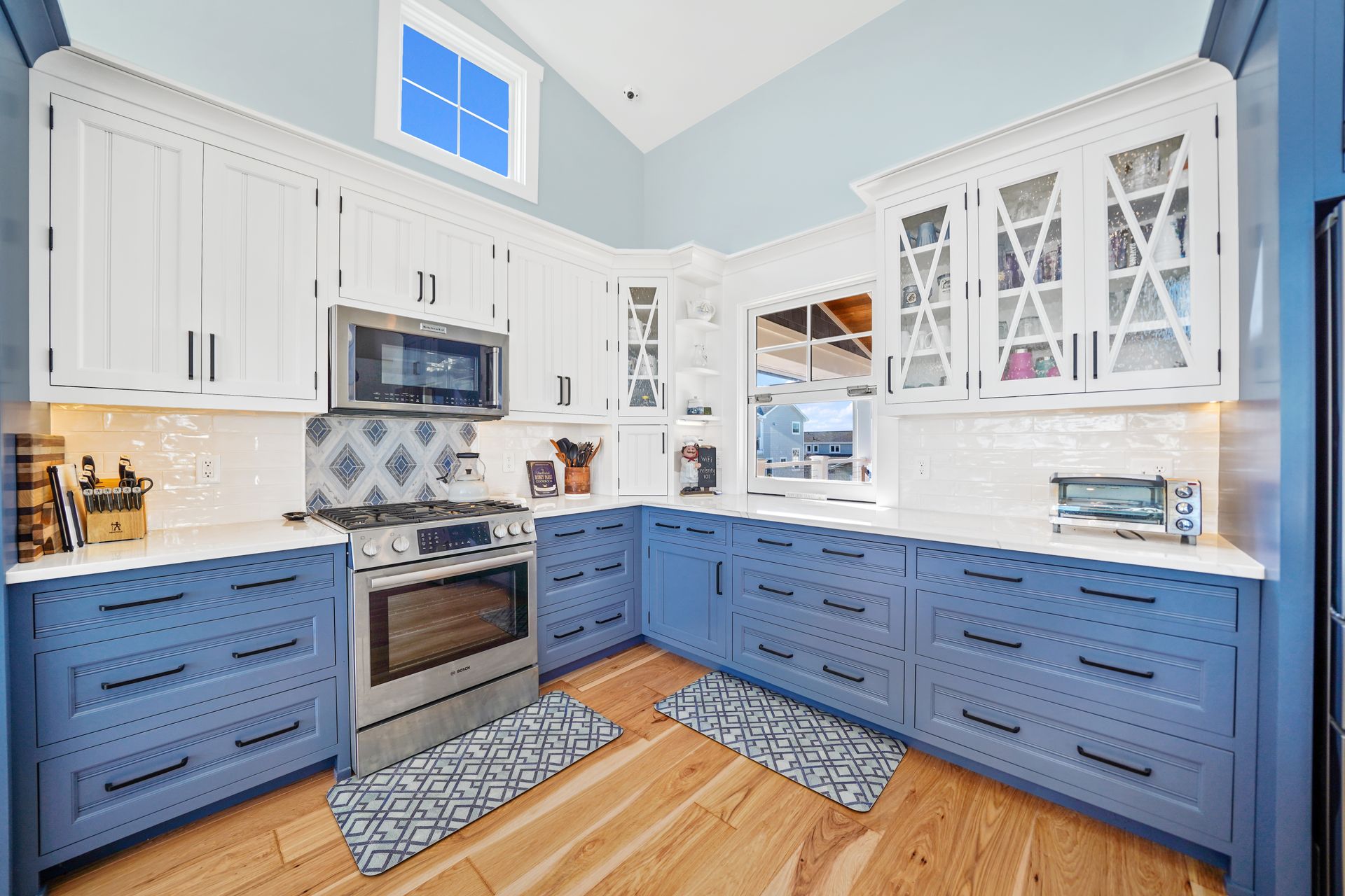 A modern kitchen with white upper cabinets, blue lower cabinets, stainless steel appliances, and wood floors.
