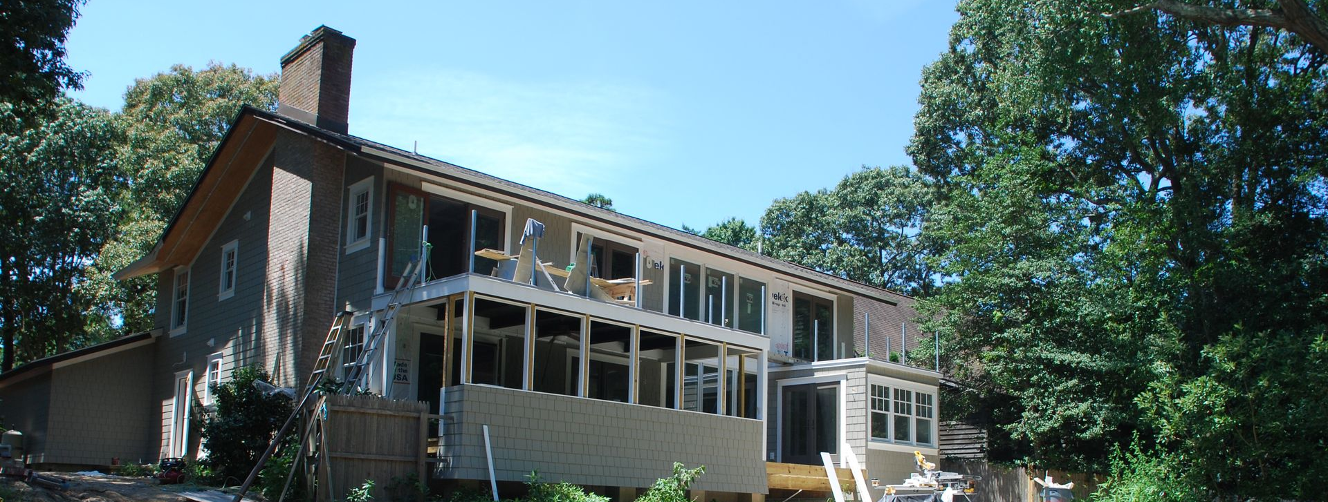 Two-story gray house with screened porch and chimney, surrounded by trees on a sunny day