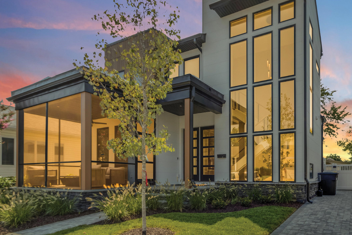 Modern two-story house with large windows and warm interior lights at dusk, framed by a small front tree and lawn