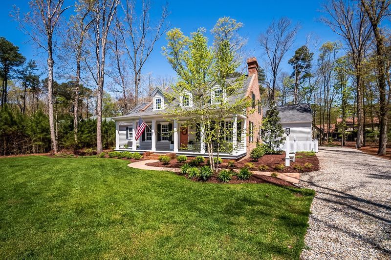 Colonial-style house with green lawn, covered porch, and gravel driveway on a sunny day.