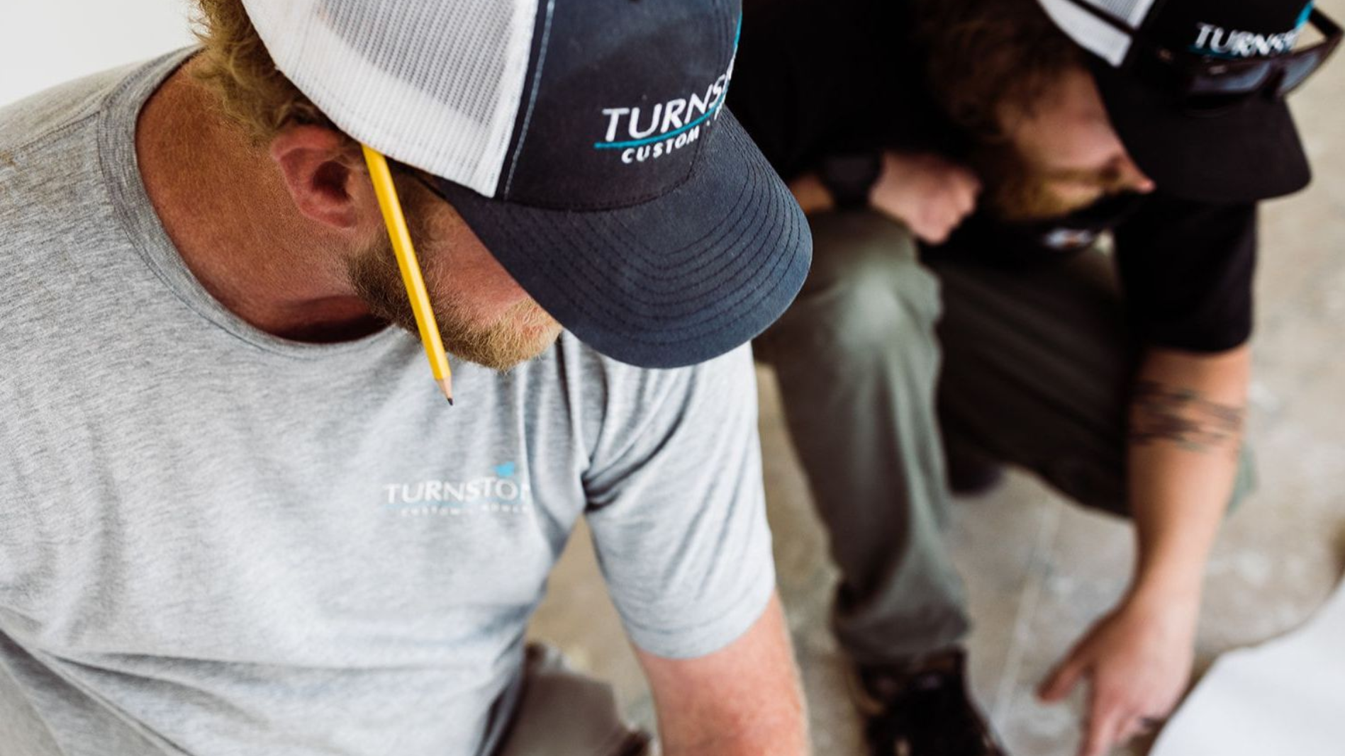 Two workers in gray shirts and black caps leaning over a task indoors with a pencil behind one ear