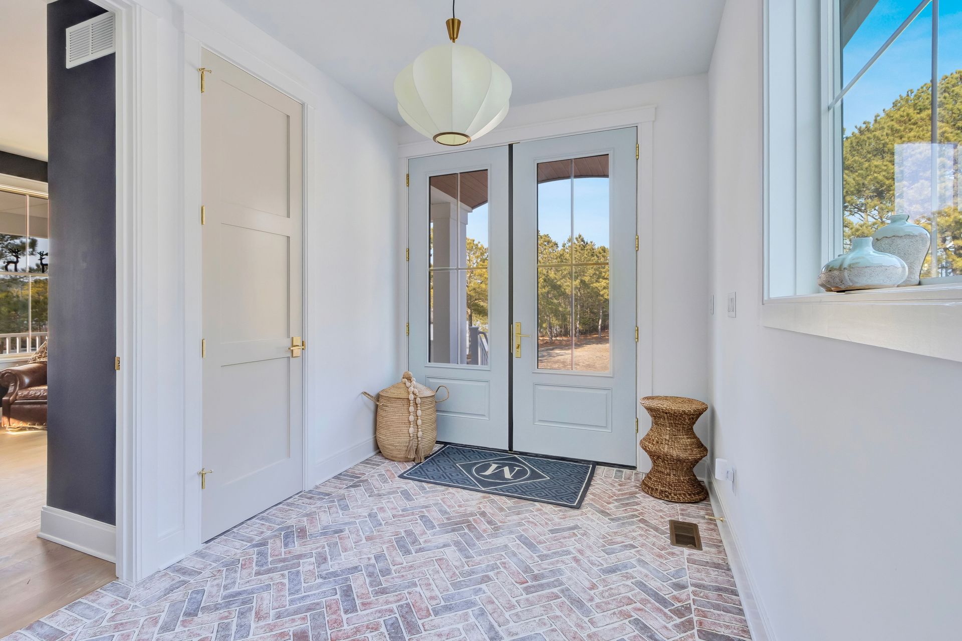Entryway with double doors, patterned floor, light fixture, side table, woven basket, and a window overlooking greenery.