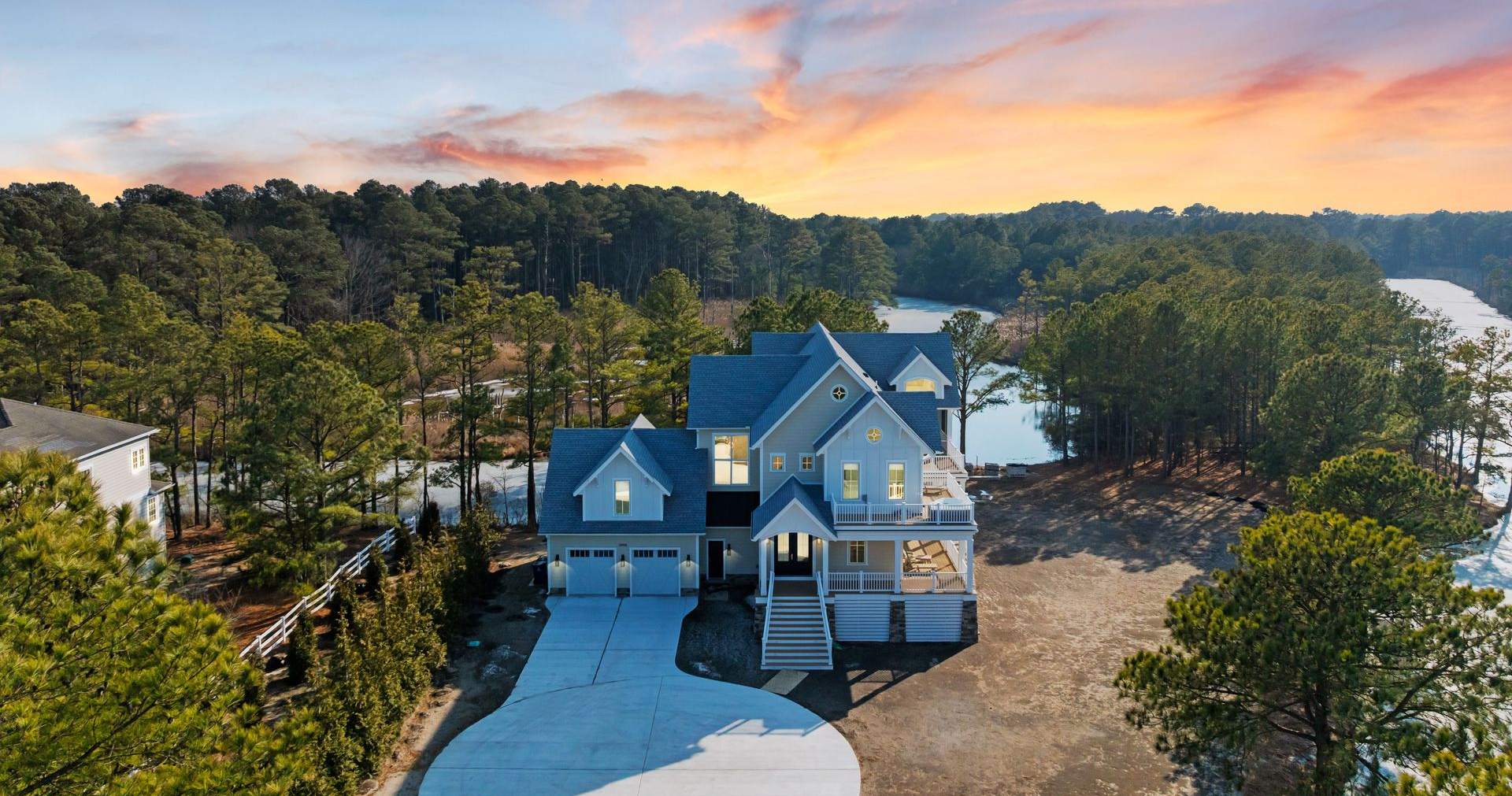 An aerial view of a large, modern multi-story house with a white garage and porch, situated by a lake and dense trees.