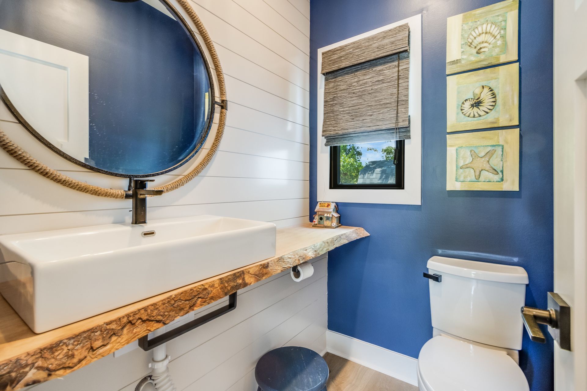 Bathroom with blue walls, white sink, wood counter, round mirror, and seashell artwork.