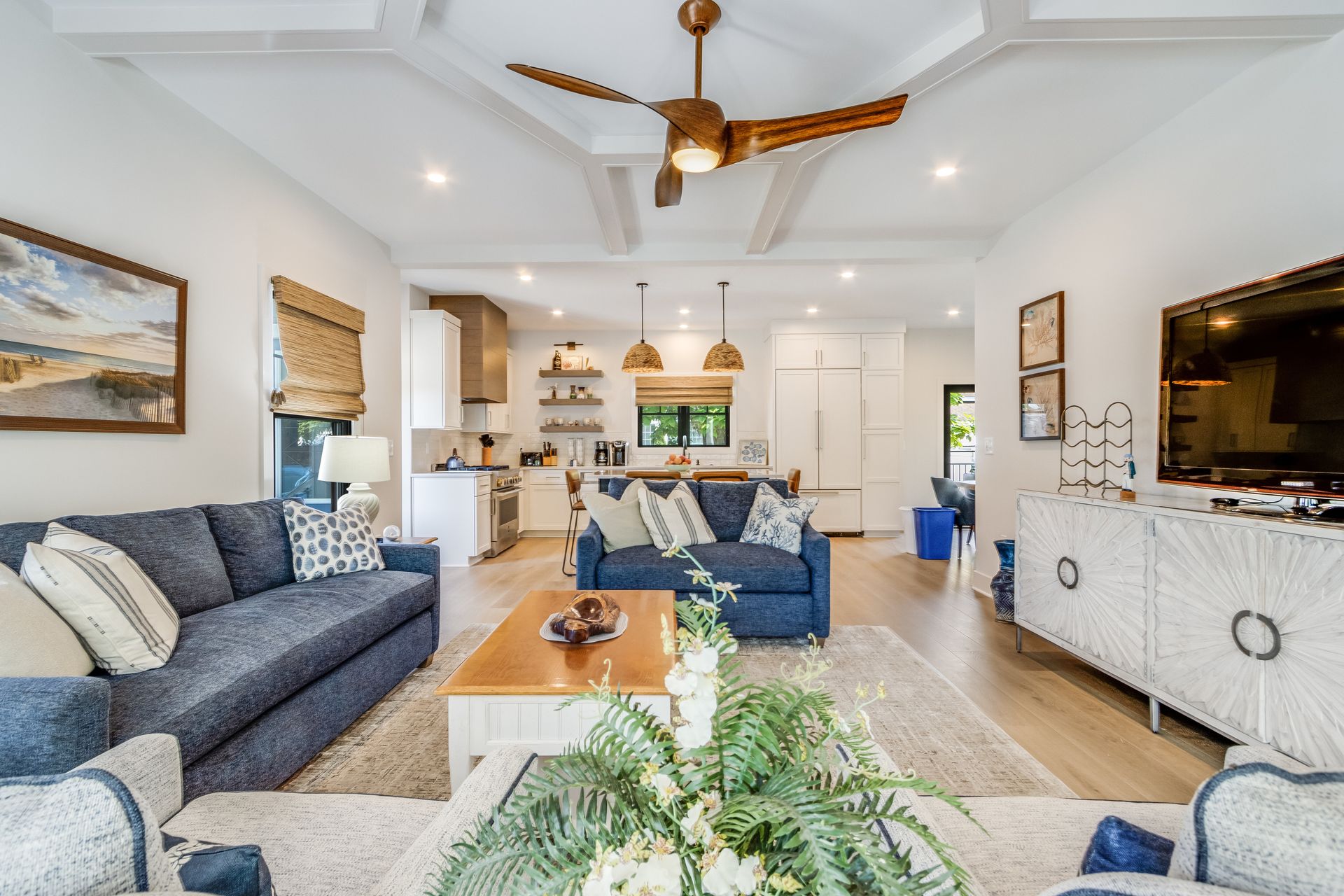 Spacious living room with blue sofas, open to kitchen. Neutral tones, ceiling fan, and white cabinets.