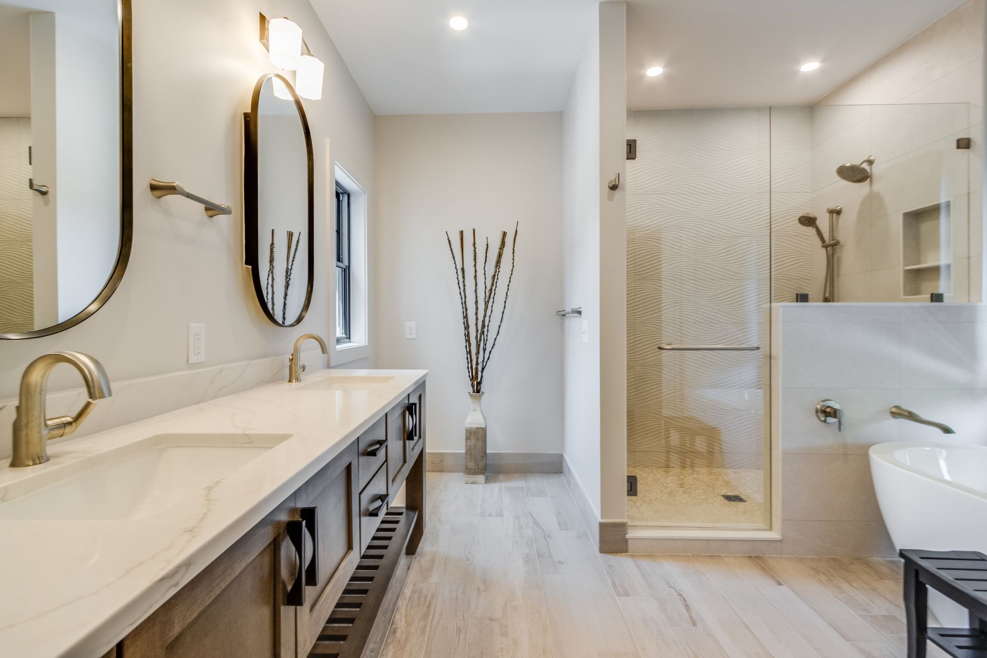 Modern bathroom with dual sinks, shower, and a freestanding tub. Light wood floors and neutral walls.