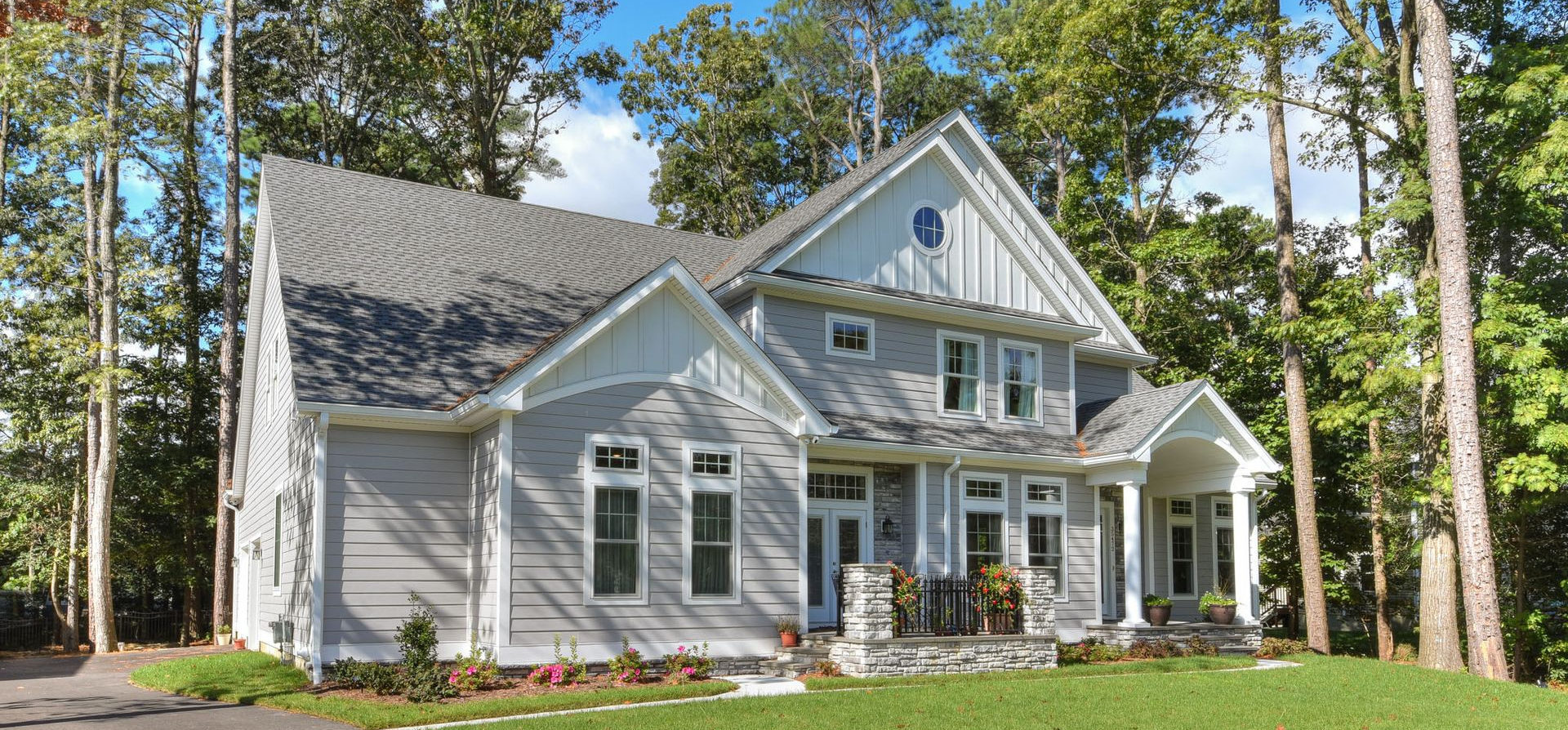 White two-story house with gray roof, front porch, and tall pine trees in a sunny yard